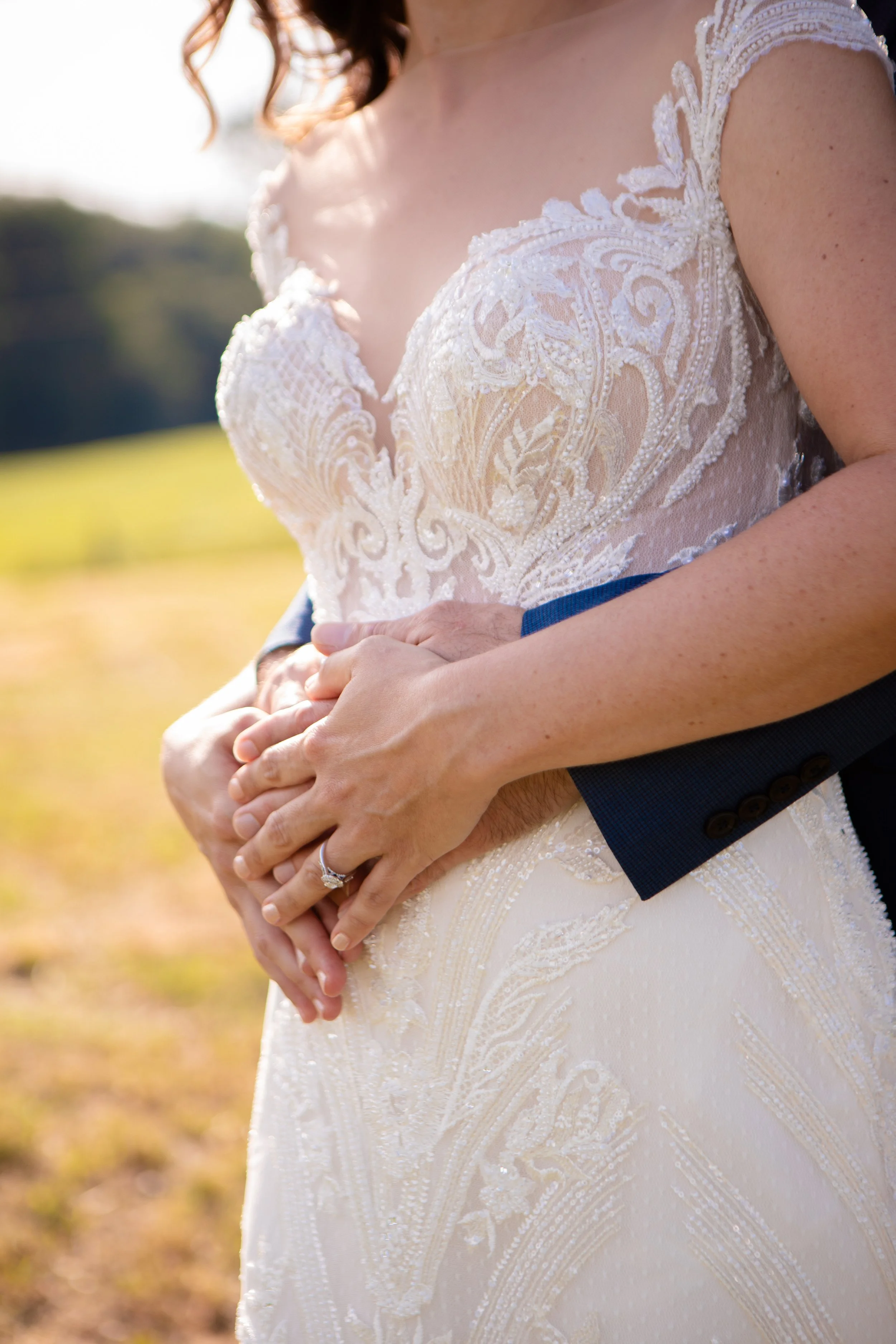 A bride in an elegant lace wedding dress is being embraced by a groom in a dark suit. The focus is on the upper body and hands, with the bride's hands resting on top of the groom's. The background is outdoors with a blurry landscape and sunlight.