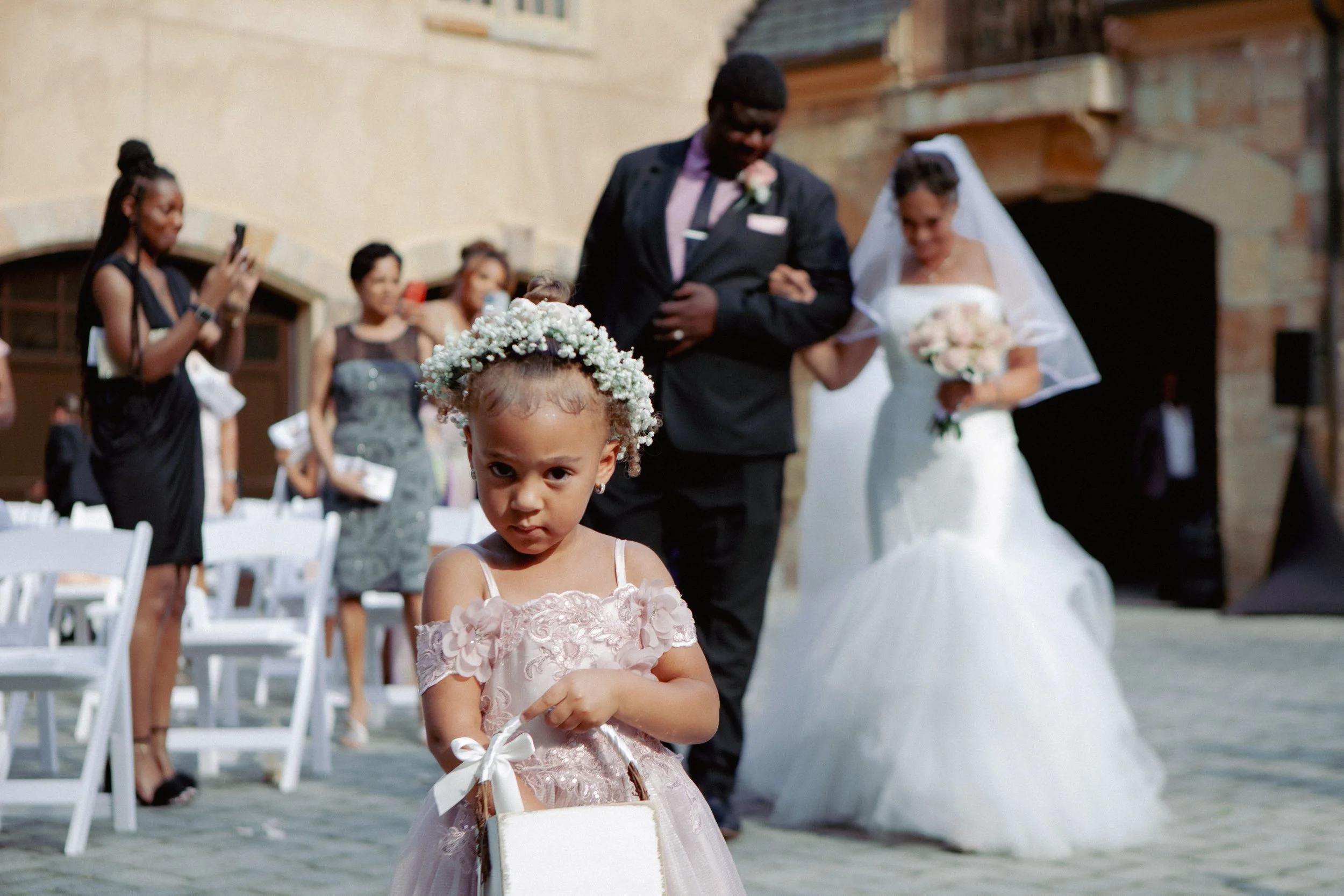A young girl with a floral wreath on her head and a pink dress holding a small basket, with a wedding ceremony in the background including a bride, groom, and guests.