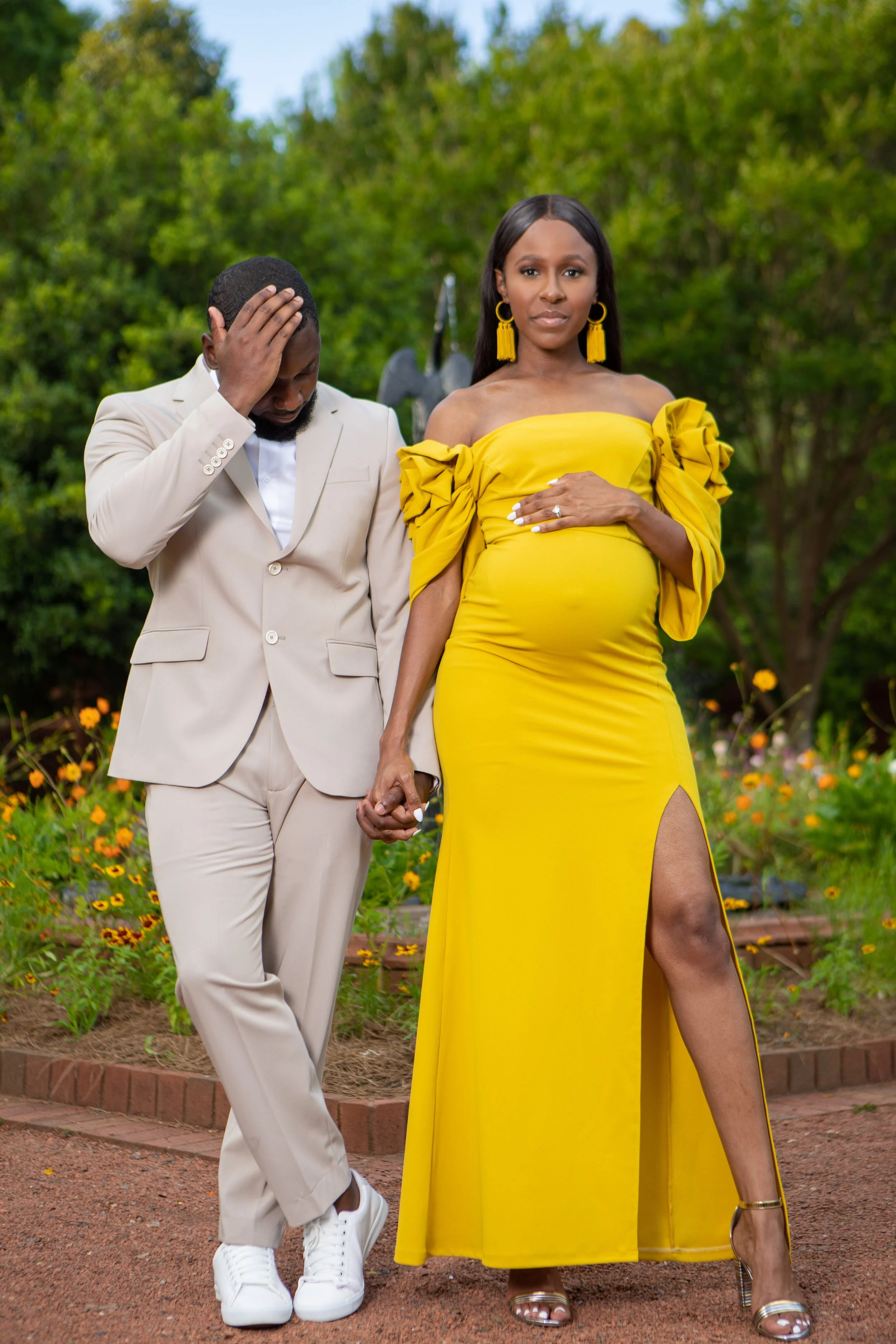 A pregnant woman in a yellow dress standing outdoors holding hands with a man in a beige suit, with lush green trees in the background.