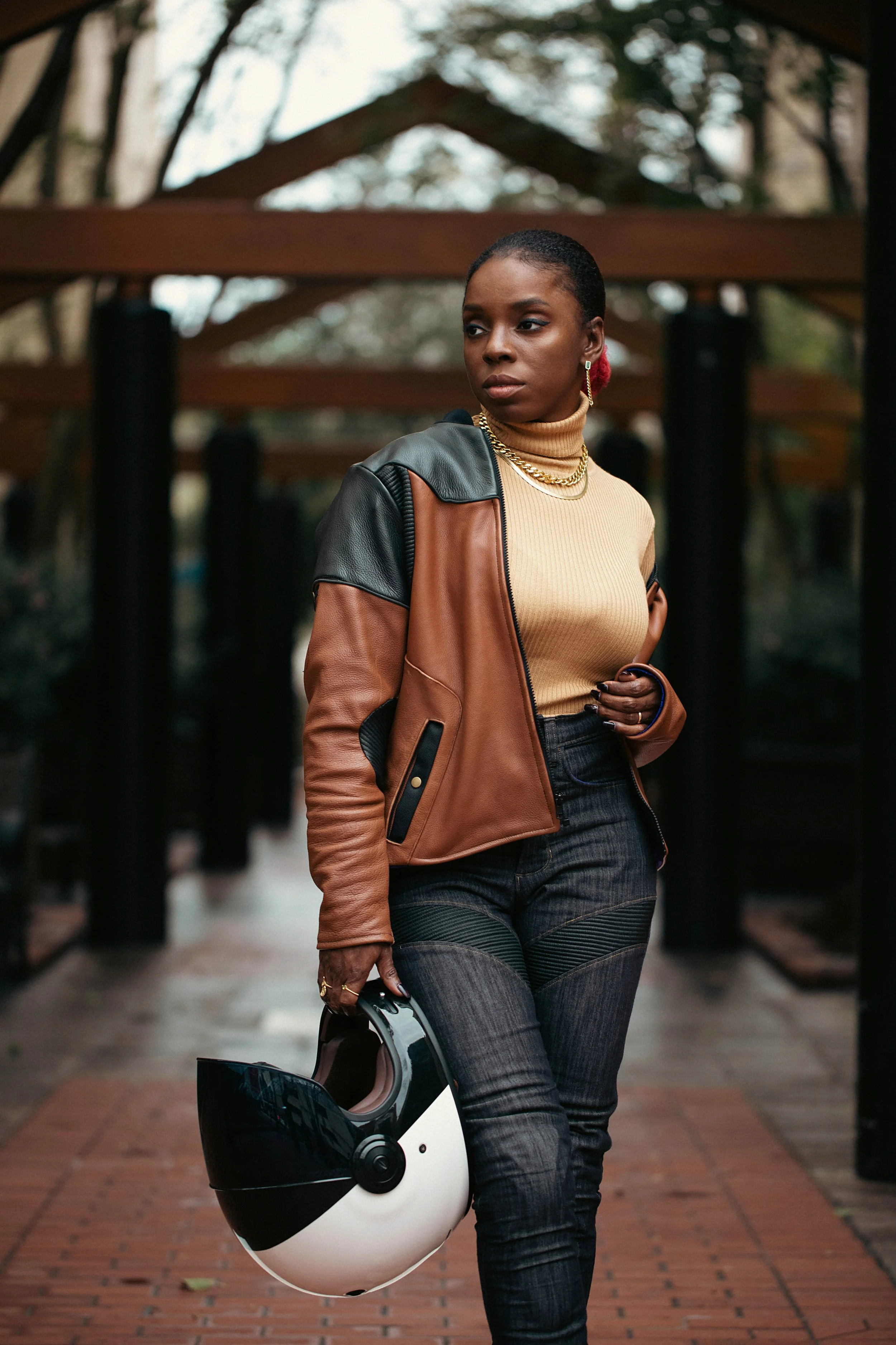 A woman walking outdoors holding a black and white motorcycle helmet, wearing a brown and black leather jacket, a beige turtleneck sweater, and black jeans, with trees and a wooden speaker structure in the background.