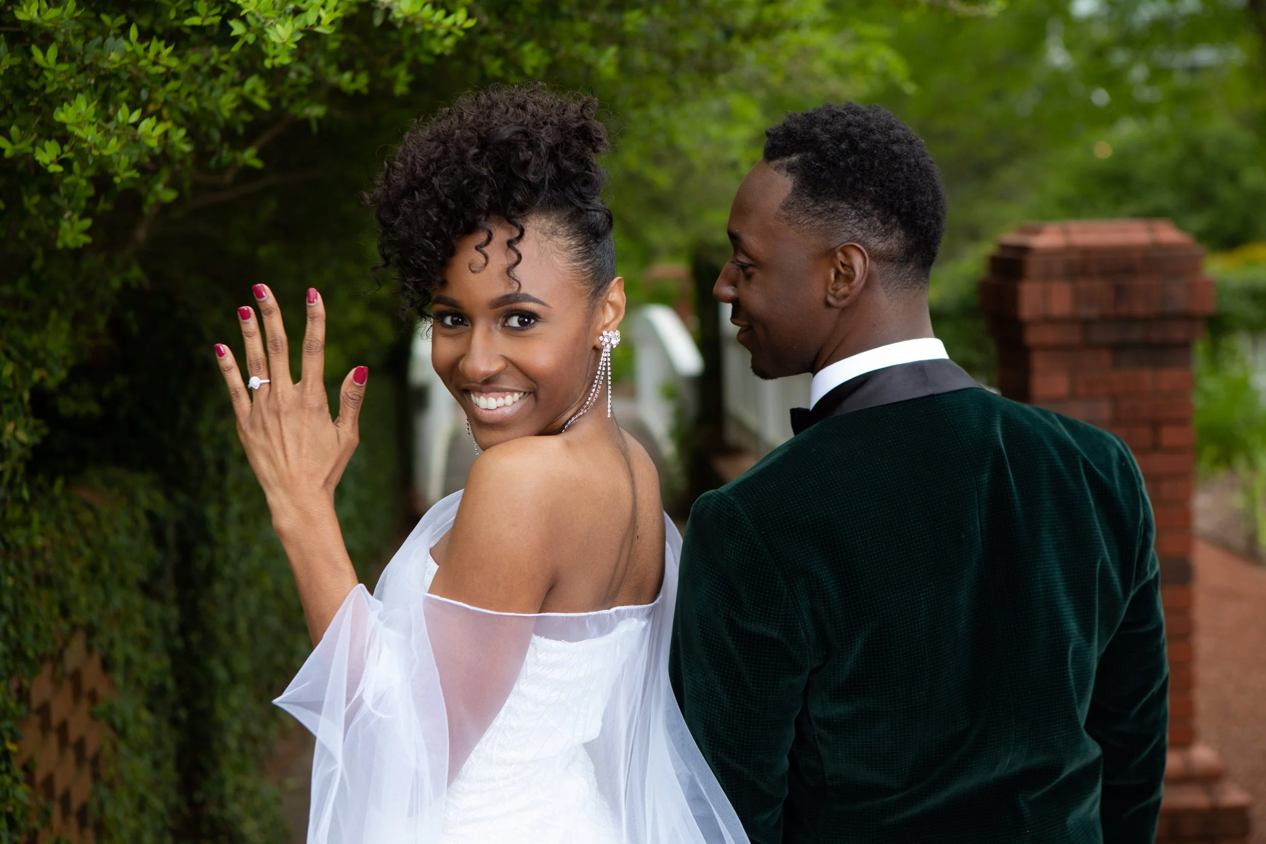 A smiling woman in a white off-the-shoulder dress showing her wedding ring, standing outdoors with greenery, as a man in a dark green tuxedo looks at her.