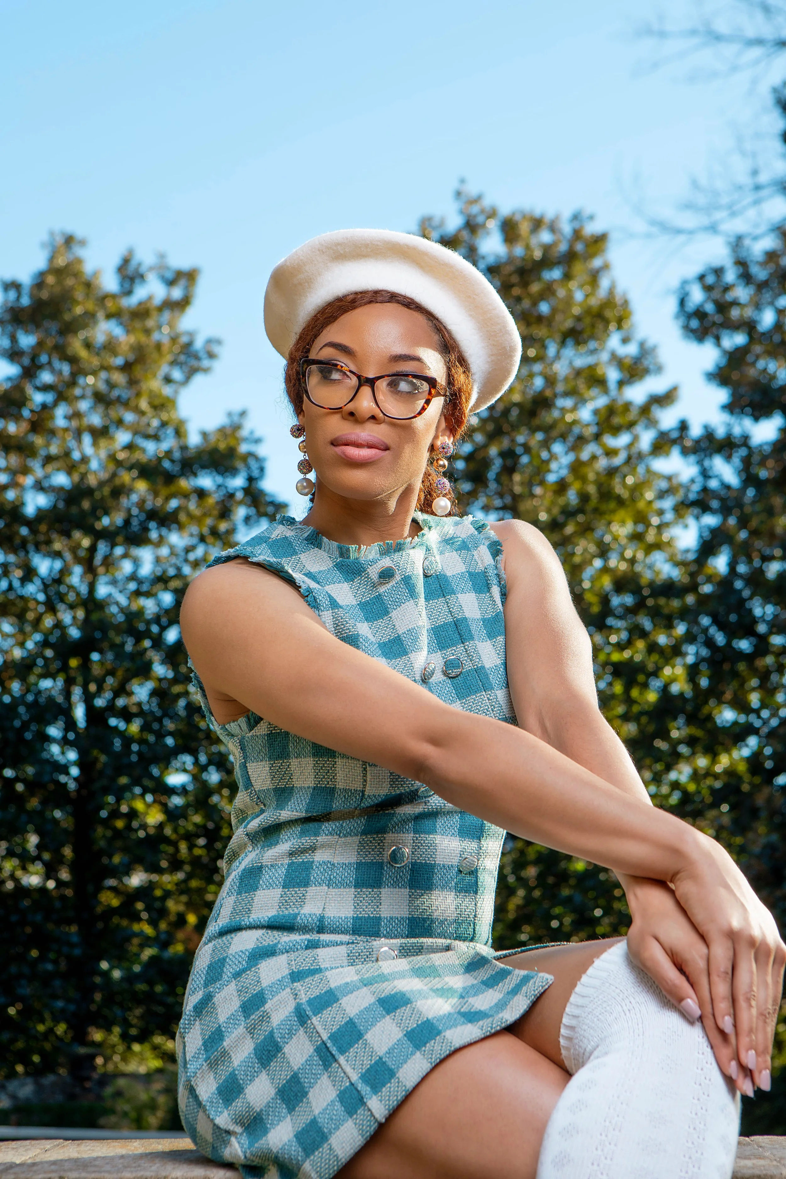 A woman wearing glasses, pearl earrings, a white beret, and a blue checkered sleeveless dress with white knee-high socks, sitting outdoors during daytime with trees and a clear blue sky in the background.