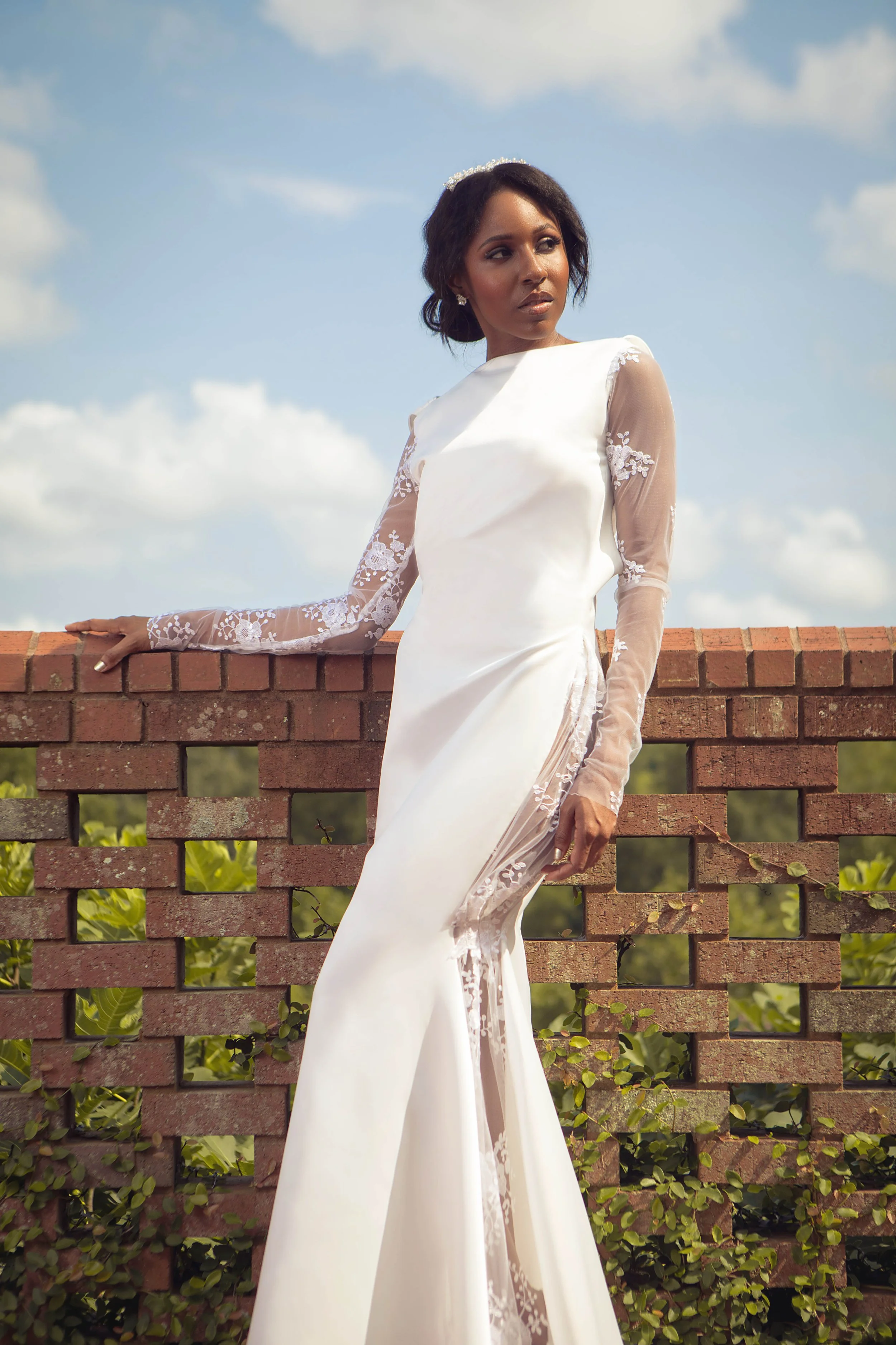 A woman in a white wedding dress with lace and sheer long sleeves stands outdoors leaning on a brick wall, with a cloudy sky in the background.