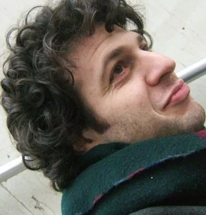 A young man with curly hair taking a selfie, smiling slightly, dressed in a dark jacket.