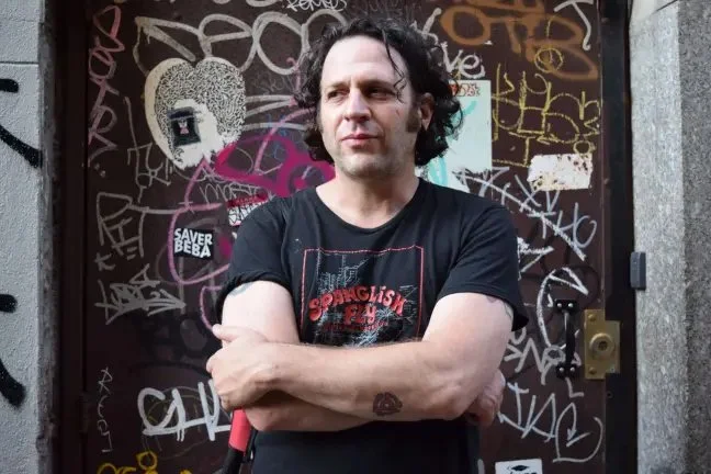 Jonathan E. Goldman standing in front of a graffiti-covered door, with arms crossed and a serious expression.