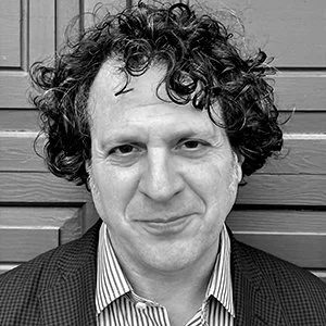 Black and white close-up of a man with curly hair, wearing a striped collared shirt and a blazer, standing against a wooden wall.