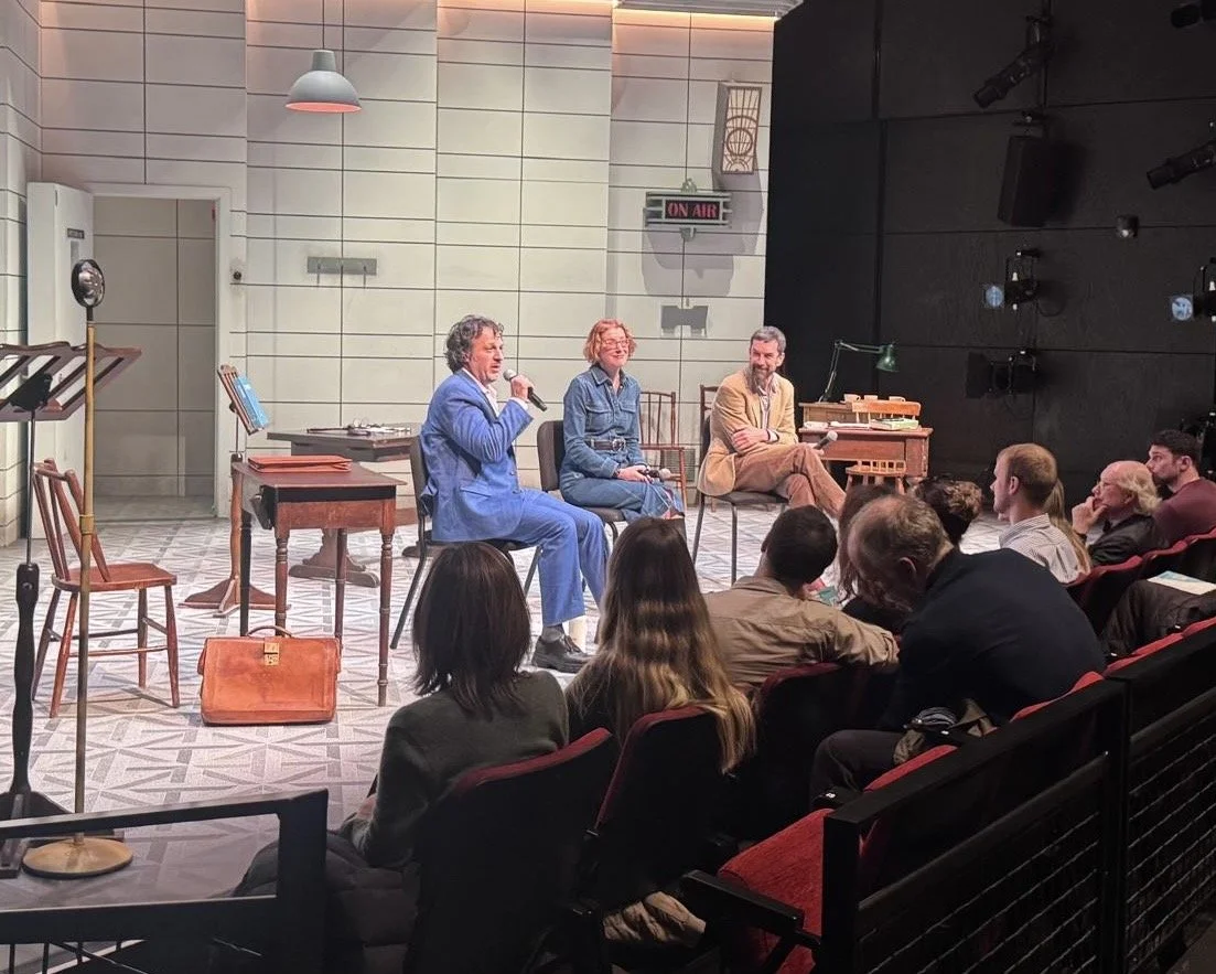 A panel discussion on a stage with Jonathan E. Goldman and two other people speaking into microphones, an audience seated facing them, and a sign that reads 'ON AIR' on the wall.