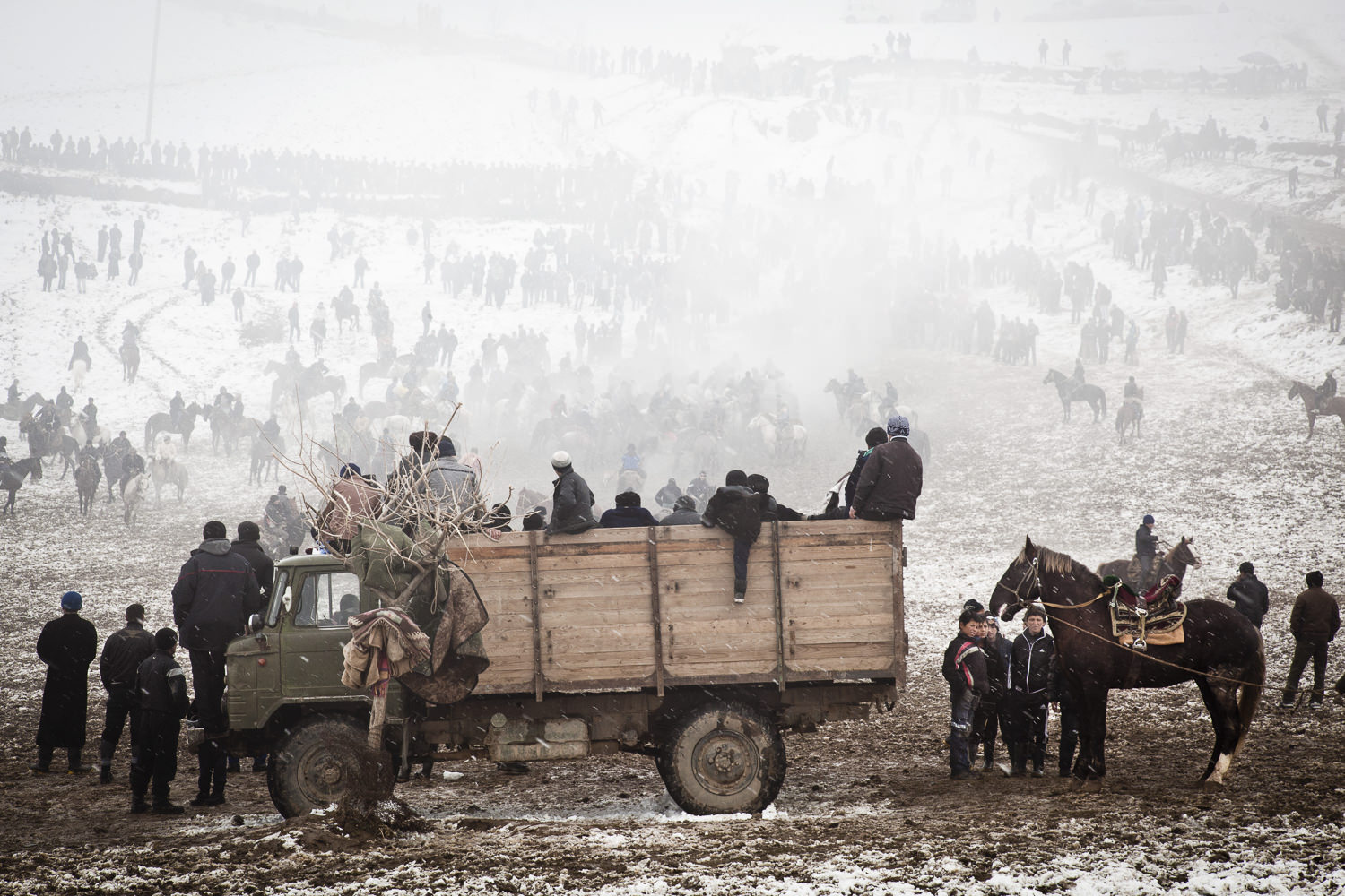  Spectators rally around a snowy buzkashi field. The truck in the foreground is both the prize truck, the announcer's truck and the holding pen for goats (buz) for the game. 