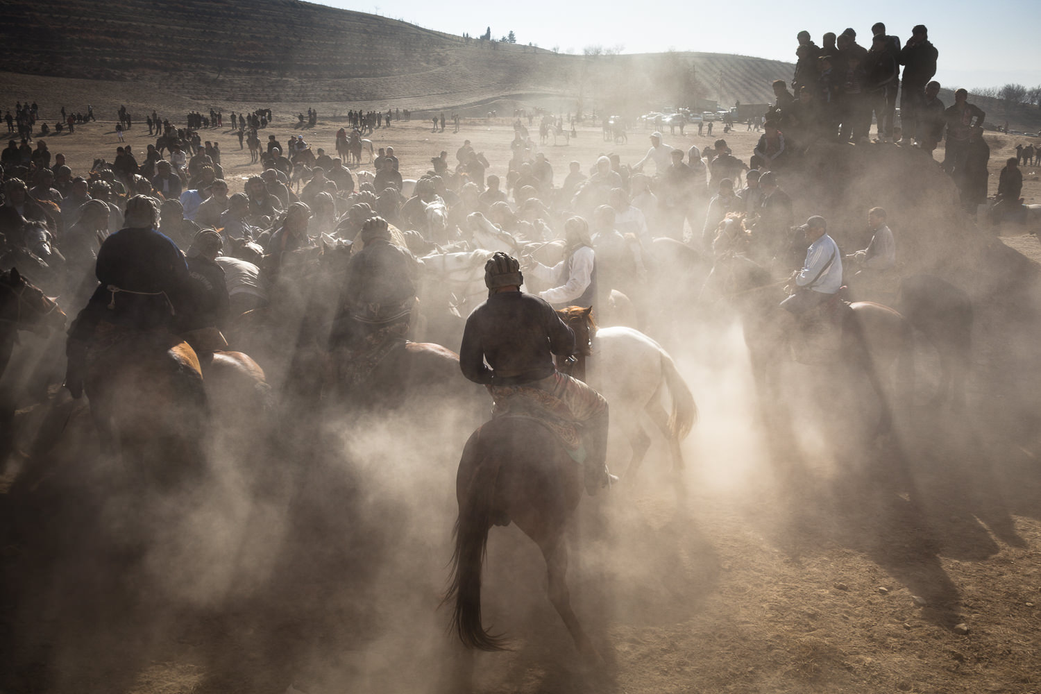  Sunlight and dust streams around Buzkashi players in Sharinav, Tajikistan. Spectators stand on a mound nearby - audience members often wander as close as possible to watch the game. 