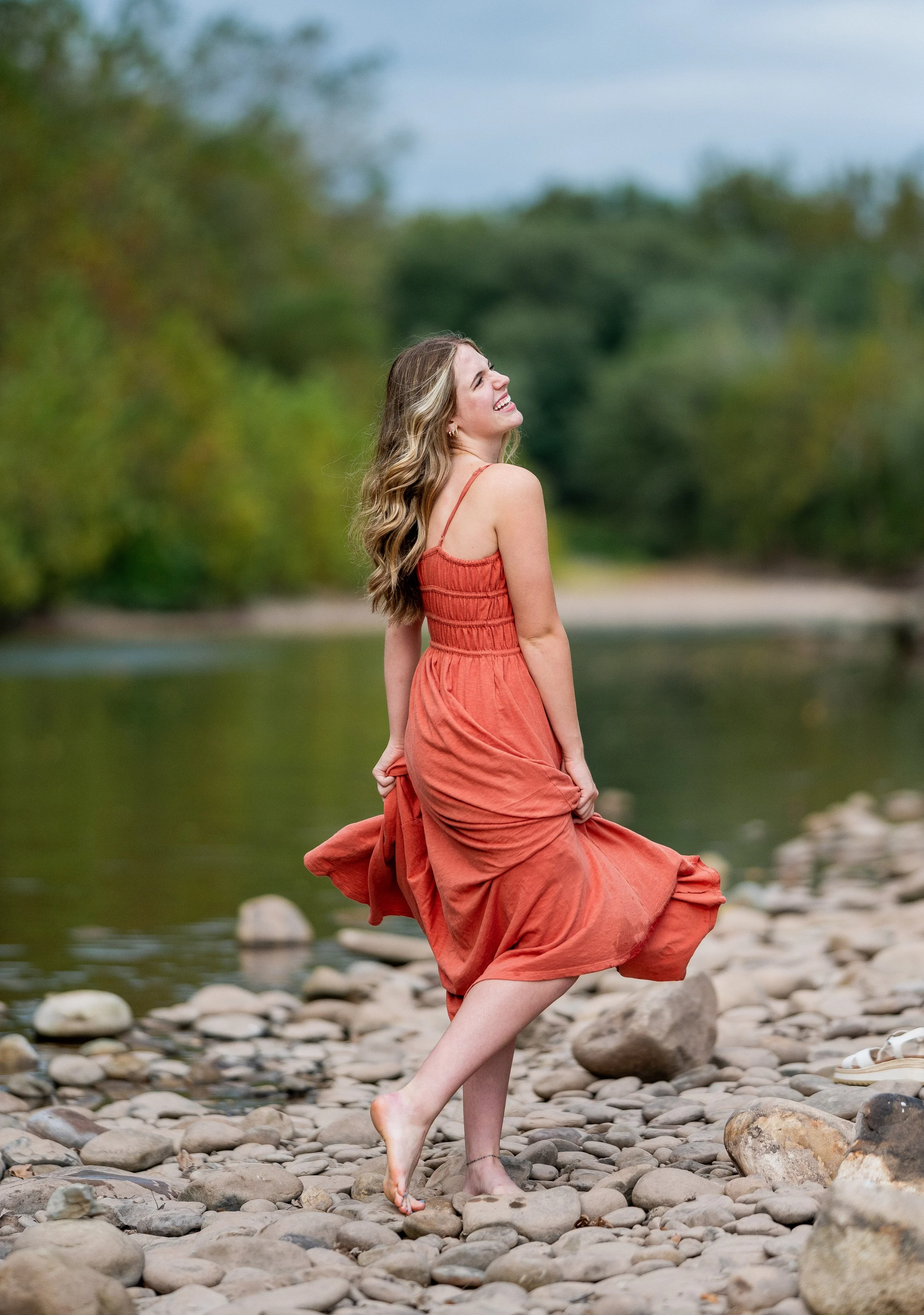 A woman in an orange dress standing on a rocky riverbank, smiling and looking up with her eyes closed, surrounded by trees.