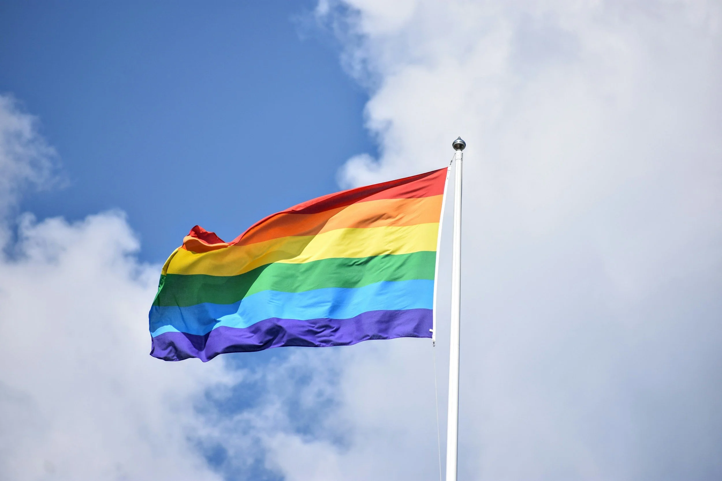 Bandeira do arco-íris hasteada ao vento contra céu com nuvens.