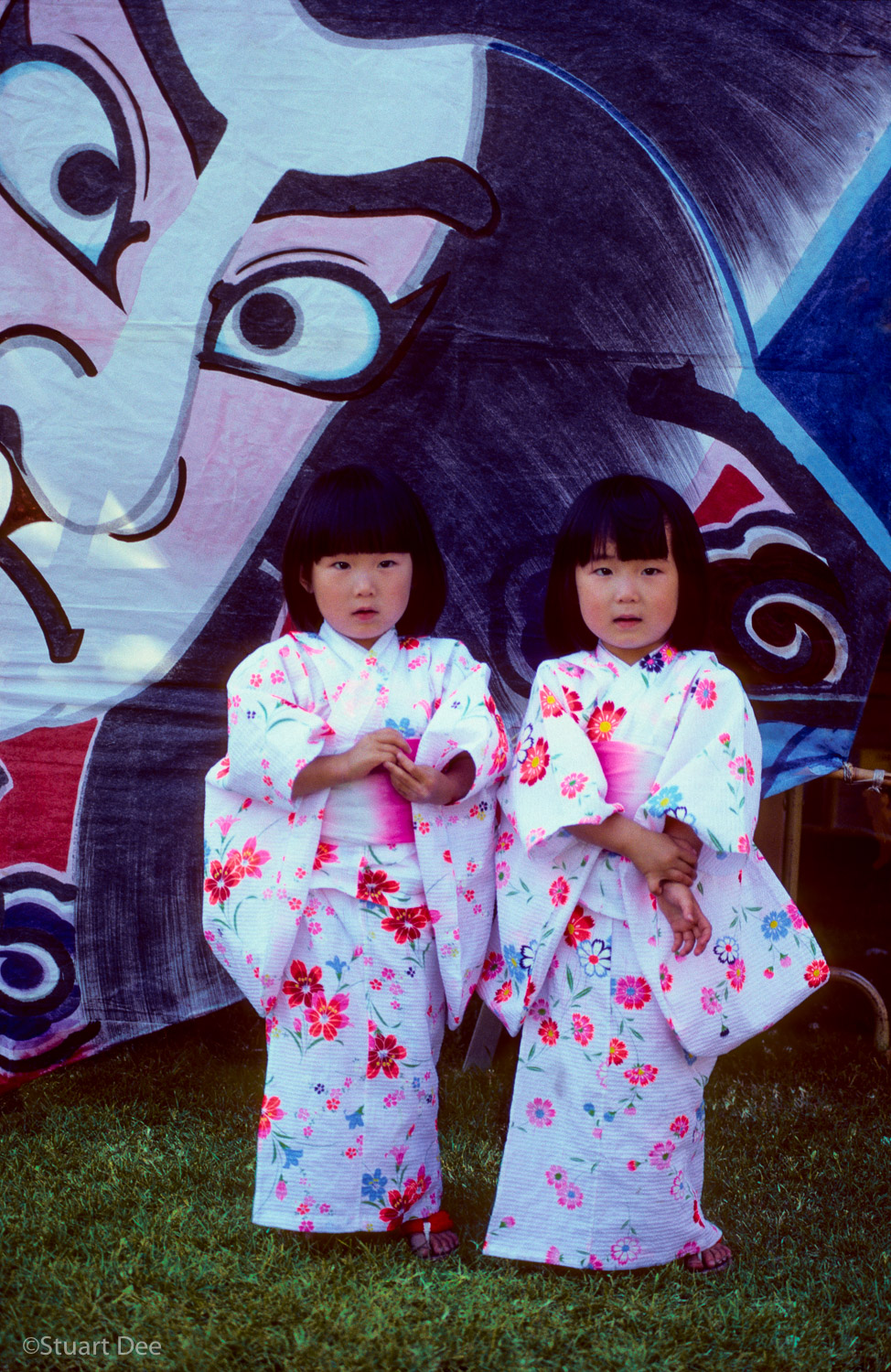  Twin Japanese girls at a Japanese festival, with a large picture of Japanese face staring at them from the background. 