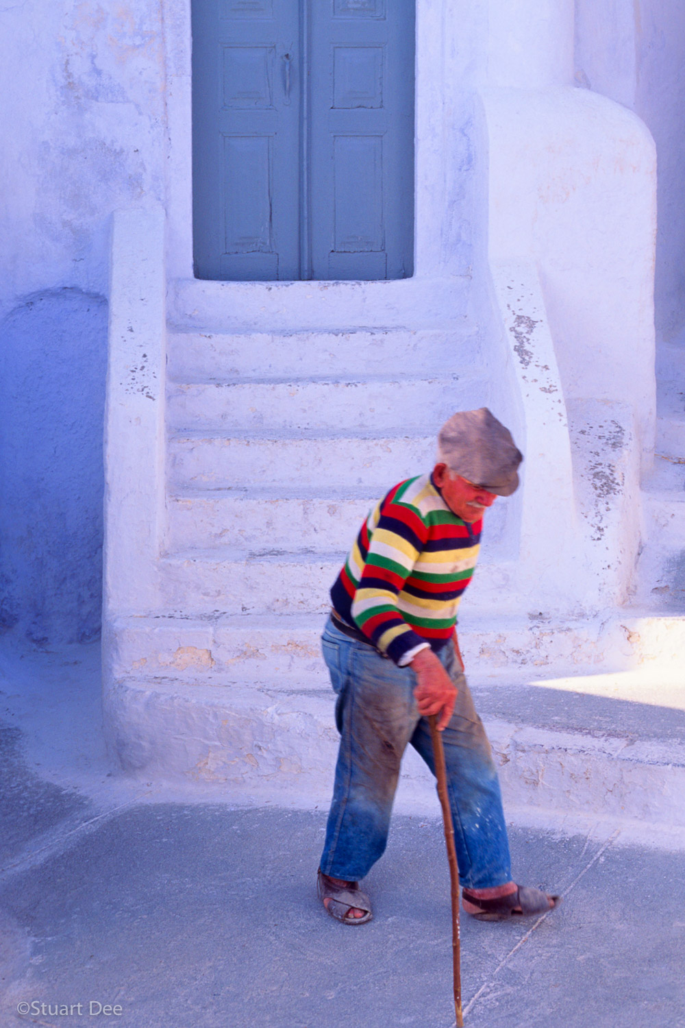 An older man walking with a cane, seen in front of a door, Pygros, Santorini, Greece 