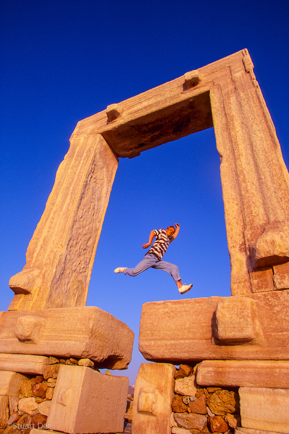  Man jumping over a gap in the marble stones of the Portara at sunset.  The symbol of Naxos, the portara is the gate of the unfinished Temple to Apollo, Naxos, Greece 