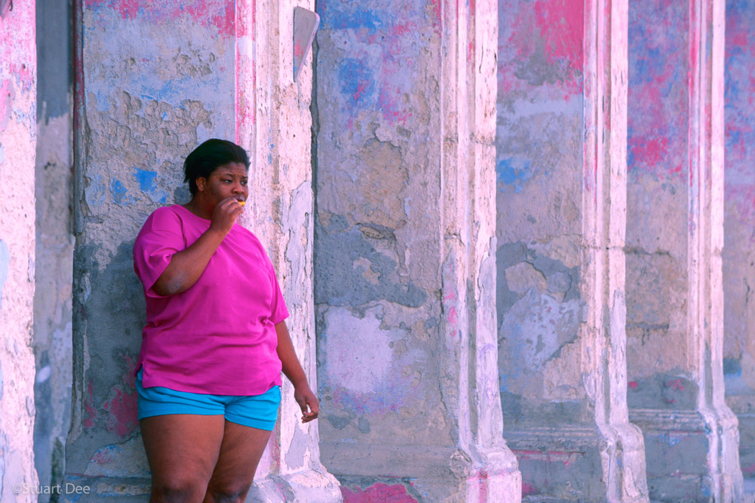  Obese woman standing among columns of an old building with chipped pink and blue paint, Malecon, Havana, Cuba 