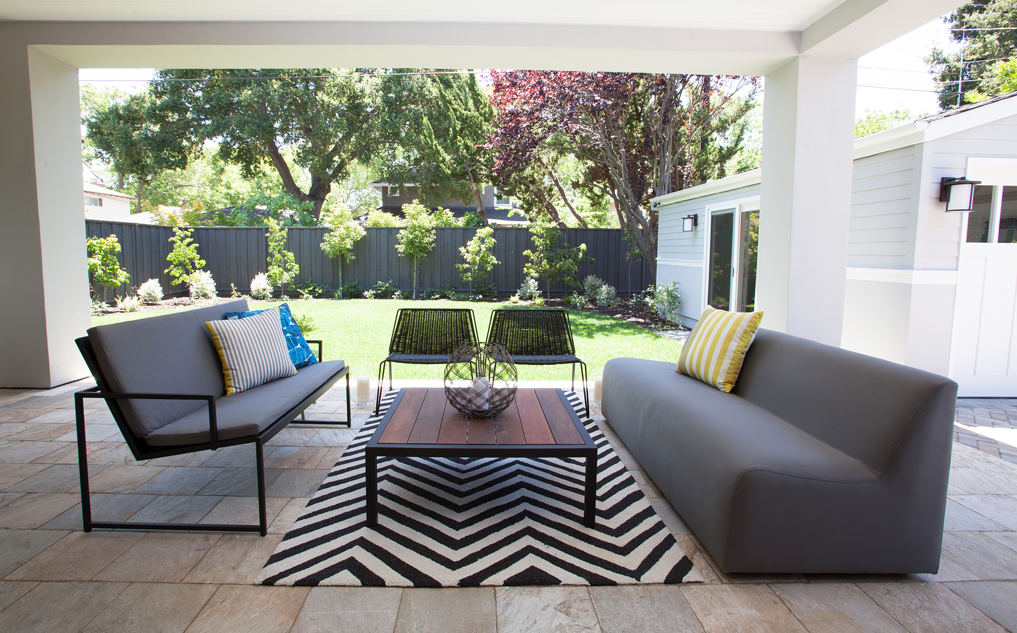 Large accordion windows from the living room open up to the outdoor patio and beautifully landscaped backyard. The seating is ample with a highlight of two modern low black chairs made of durable plastic woven across a powder coated frame. A bold black and white chevron area rug connects back to the interior.
