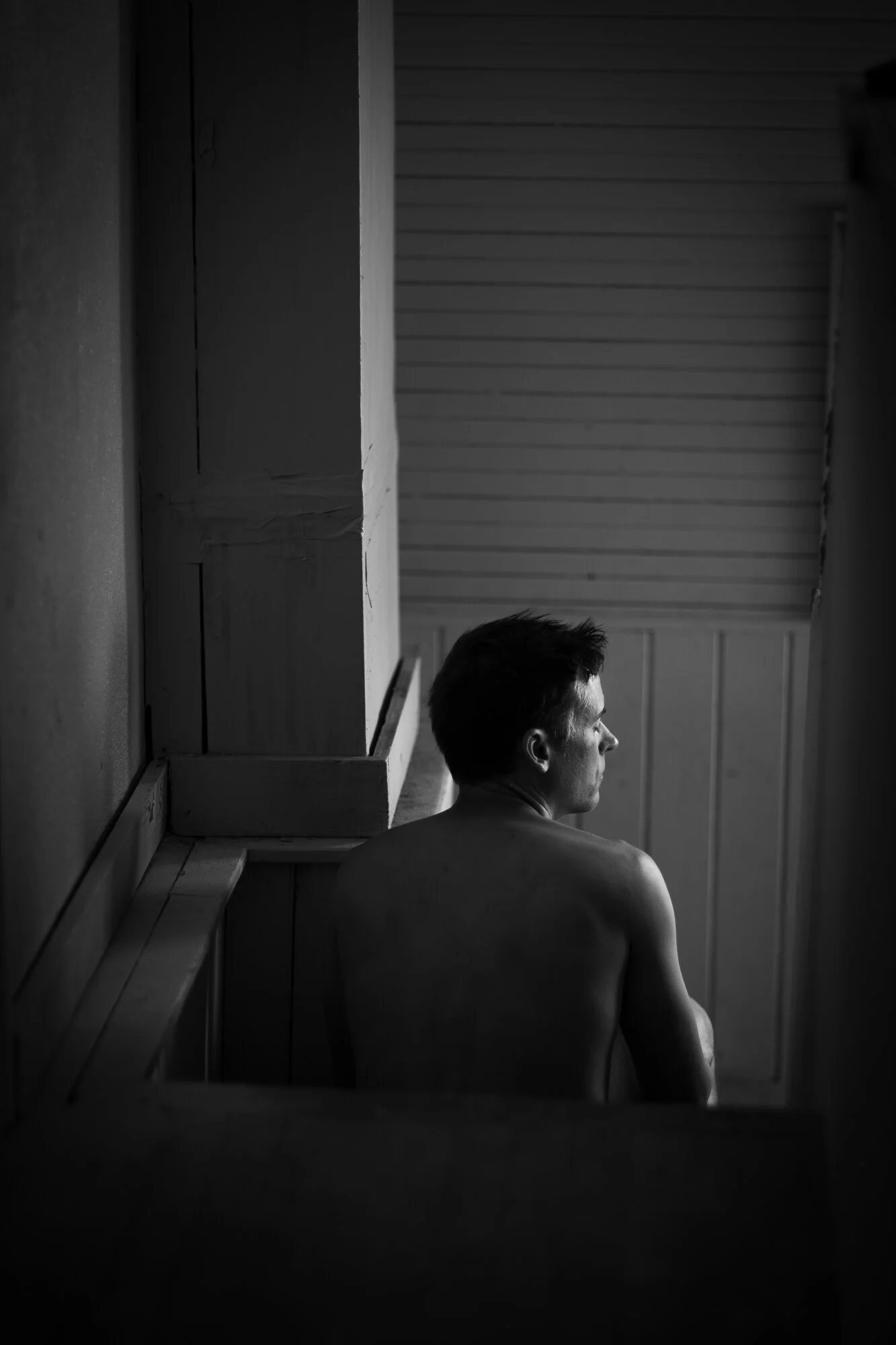 Black and white photo of a man with short hair sitting indoors, seen from the side, with his back and shoulder visible, against a wooden wall.