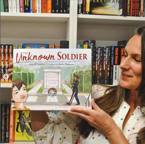Illustrator Jamie Peterson holds her book The Unknown Soldier with a backdrop of books