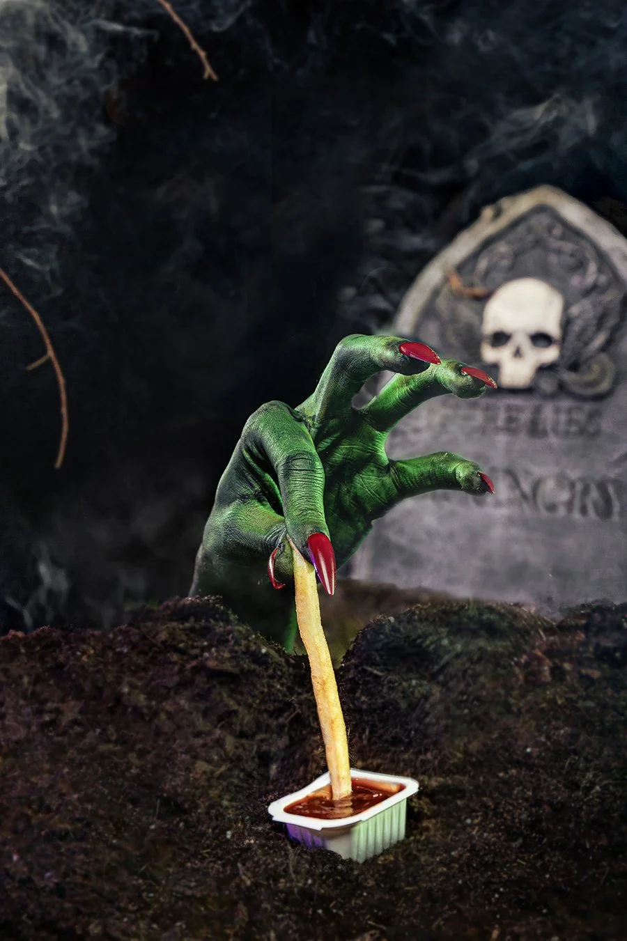 A green, zombie-like hand with red painted nails emerging from the soil, gripping a breadstick dipped in what appears to be chocolate, in front of a spooky gravestone with a skull carving.