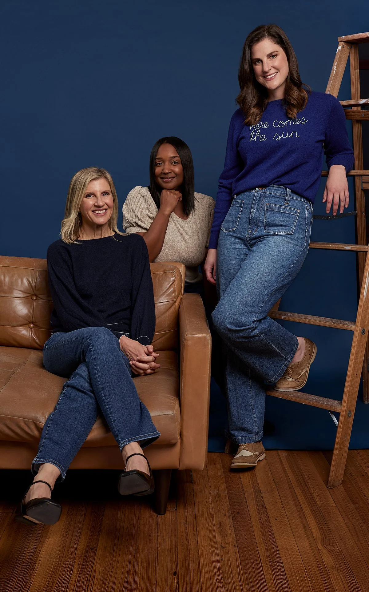 Three women posing indoors against a blue background, with a brown leather couch and a wooden ladder in the scene. The woman on the left is sitting on the couch wearing a navy sweater and jeans, smiling at the camera. The woman in the middle is stand