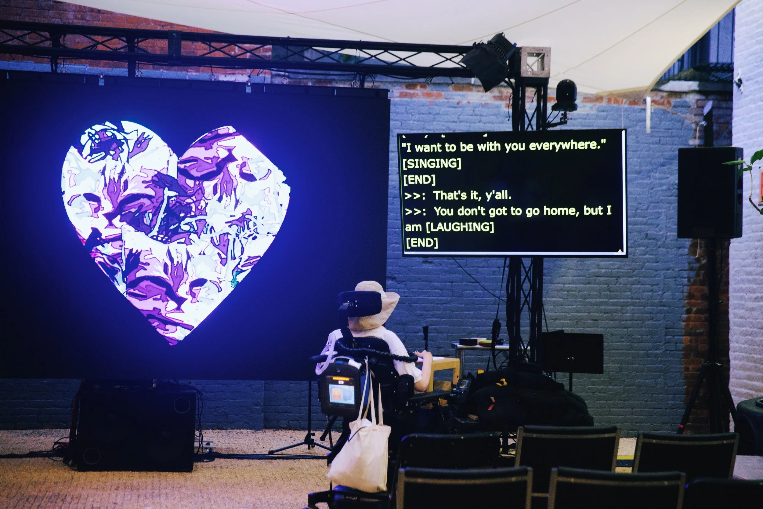  A photo by Mengwen Cao of an outdoor rehearsal space with rows of chairs and a projection screen showing a large, multicolored heart. A powerchair user in a floppy sunhat looks up at a CART monitor with captions that read “I want to be with you ever