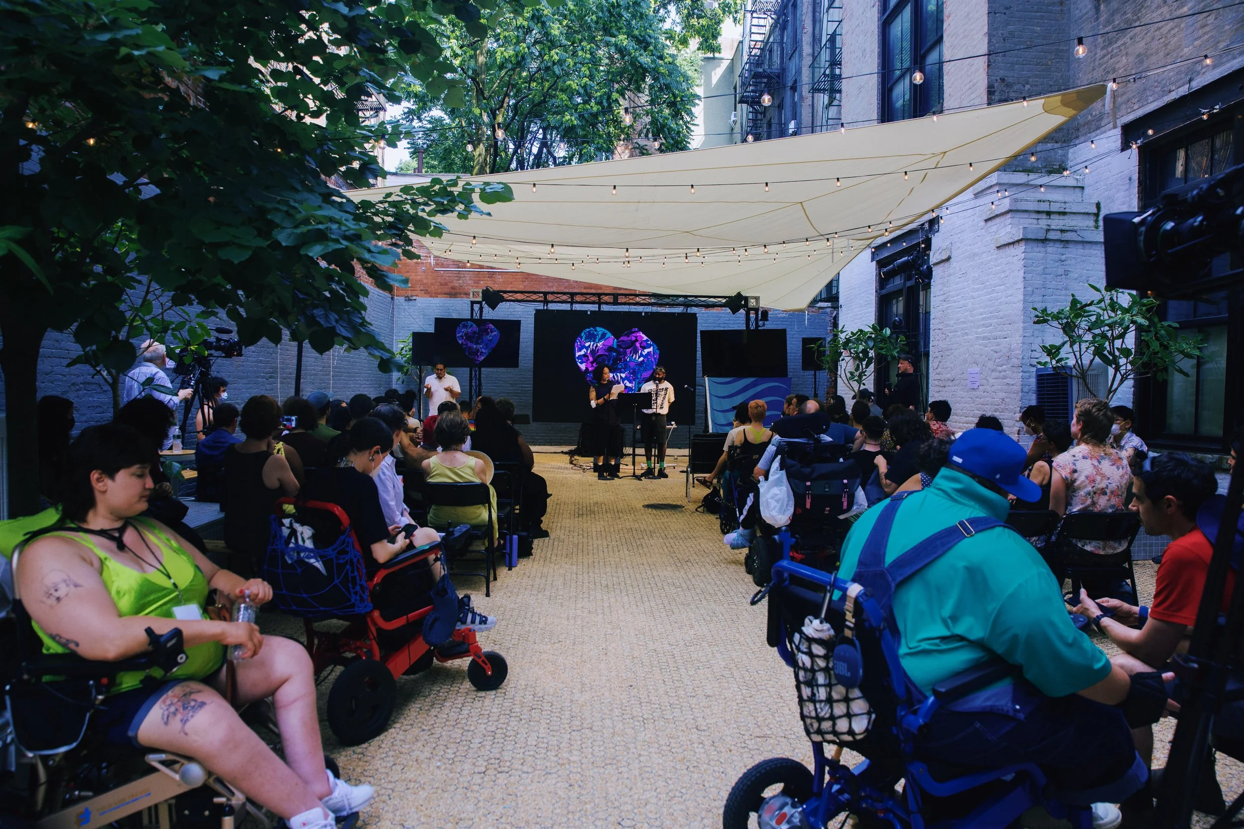  A photo by Mengwen Cao taken from the back of the Performance Space NY courtyard on a hot summer day. Under the shade of an awning and trees, a large, colorfully dressed crowd turns to face two people at a microphone. 