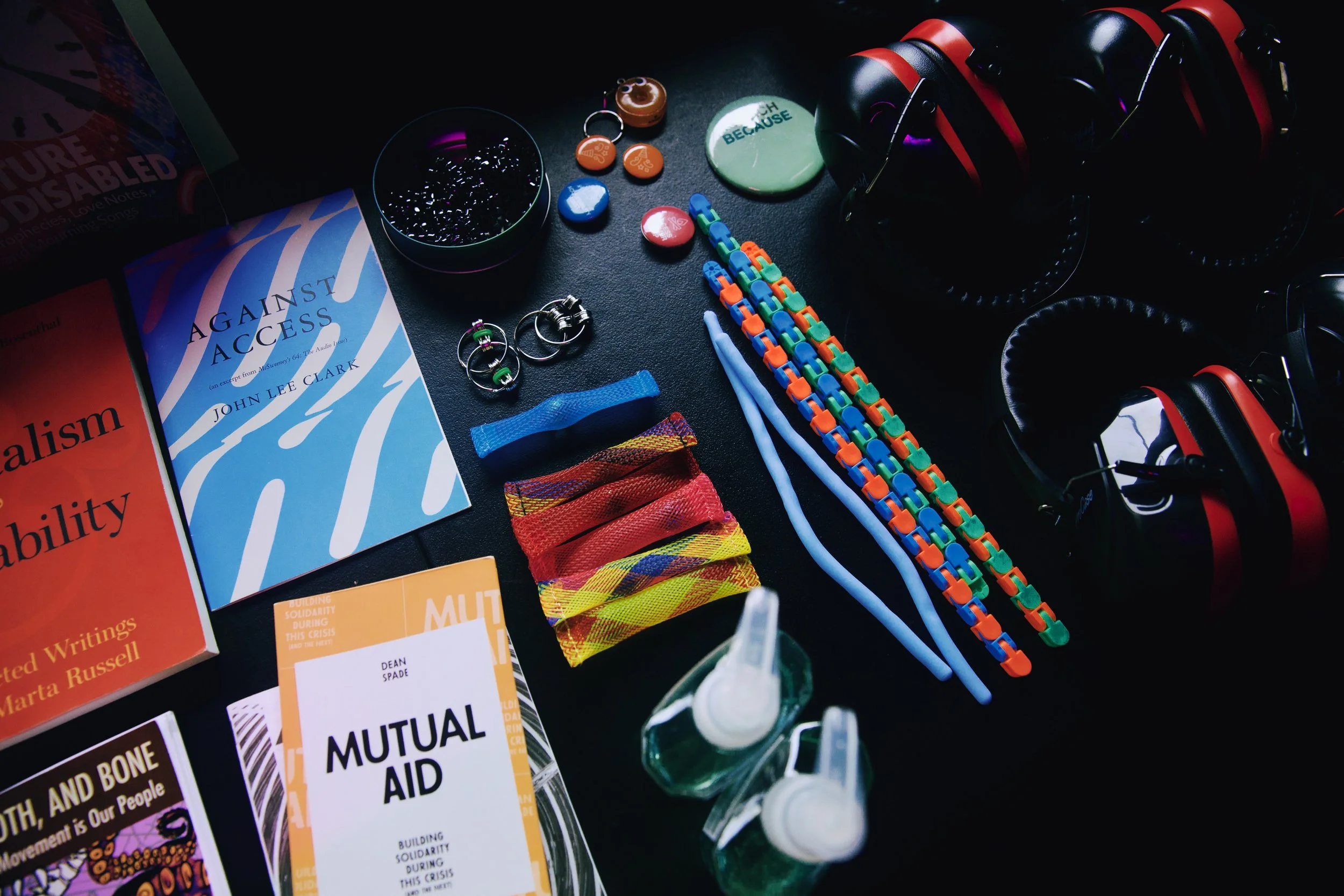  A photo by Mengwen Cao shows a table covered with stim toys, ear defenders, buttons, hand sanitizer, and books, including  Against Access  by John Lee Clark,  Mutual Aid  by Dean Spade,  Capitalism and Disability  by Marta Russell, and  Skin, Tooth,