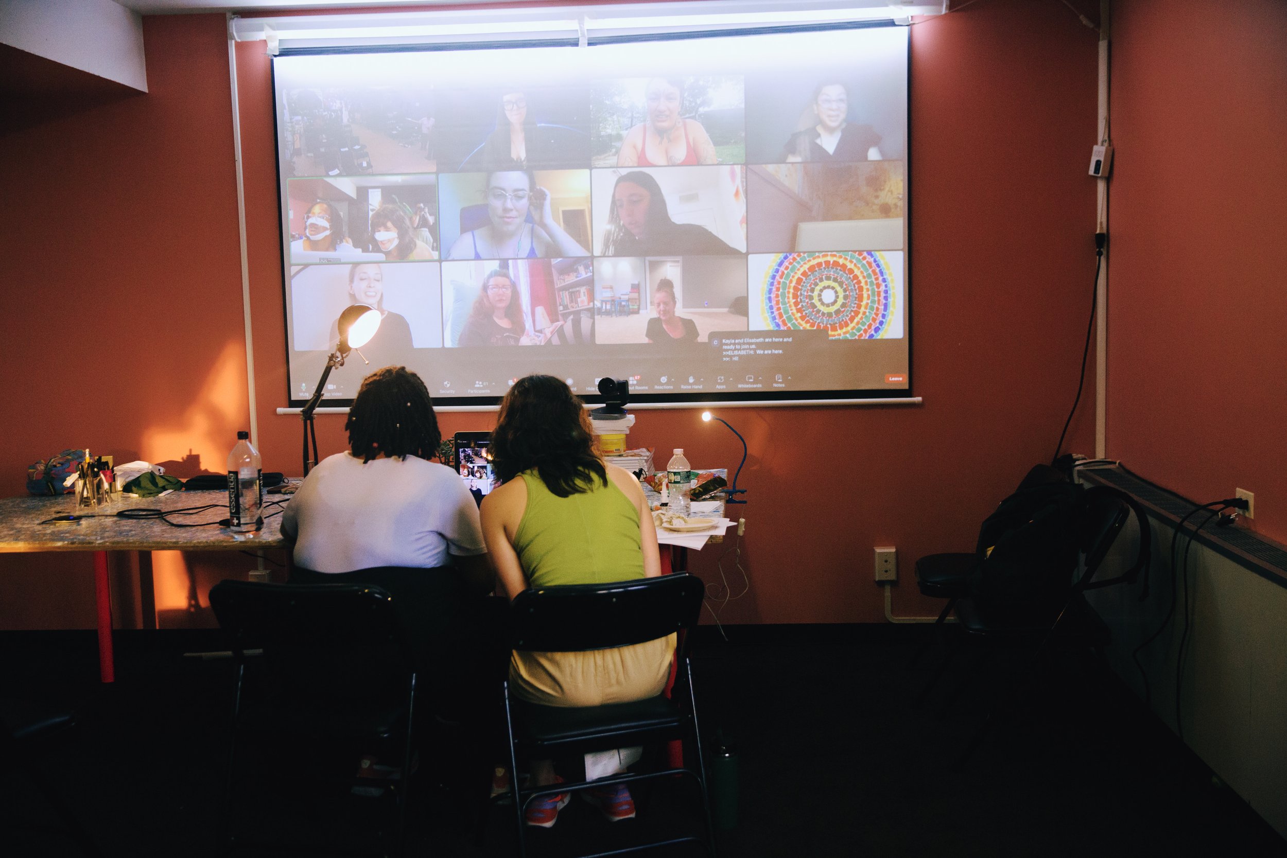  A photo by Mengwen Cao shows two people in summer clothing leaning in close together over a laptop at a cluttered table. Above them, a projection screen shows a zoom room full of people in indoor and outdoor spaces. 