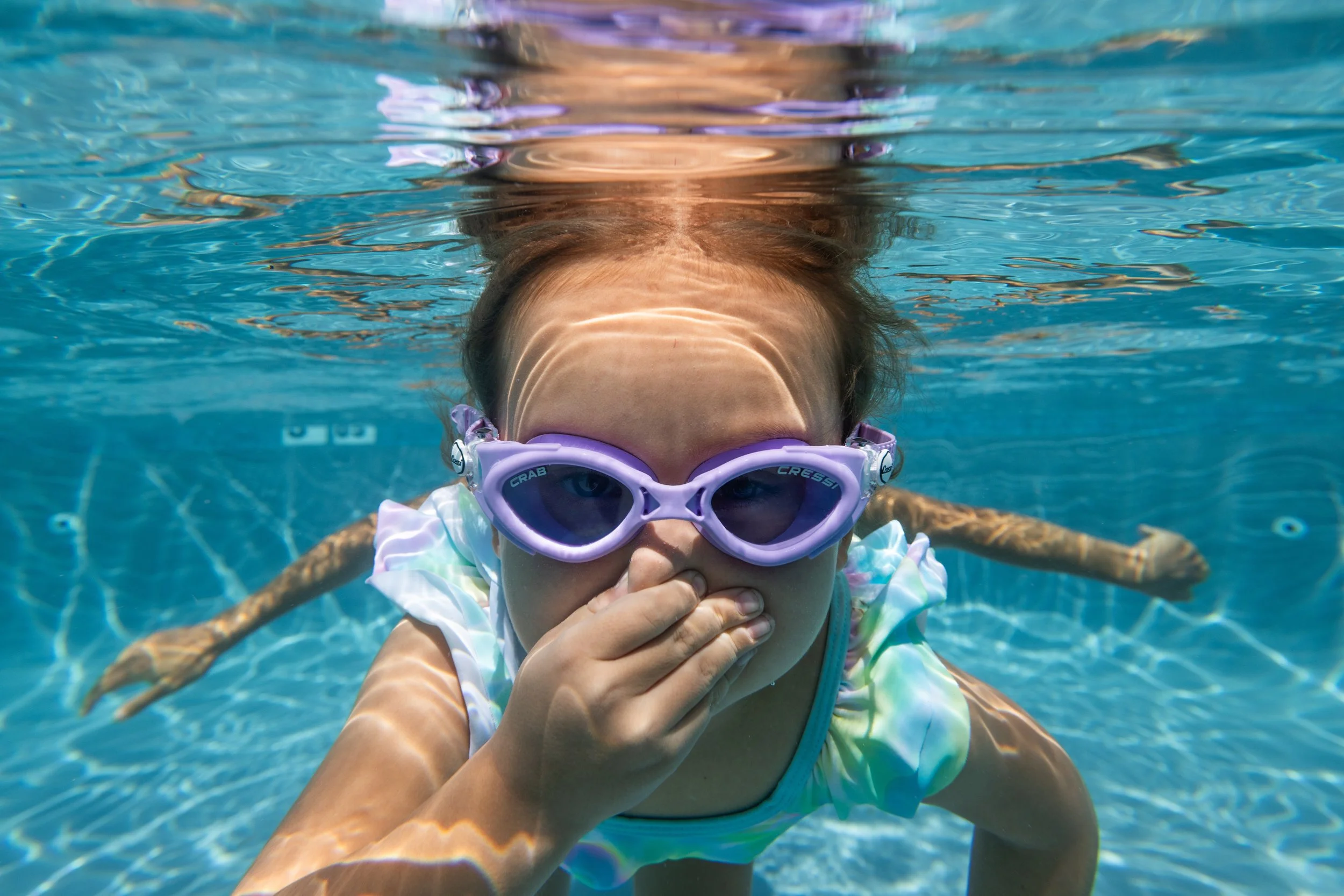 A young girl with glasses and long hair swimming underwater in a pool, covering her nose with her hand.