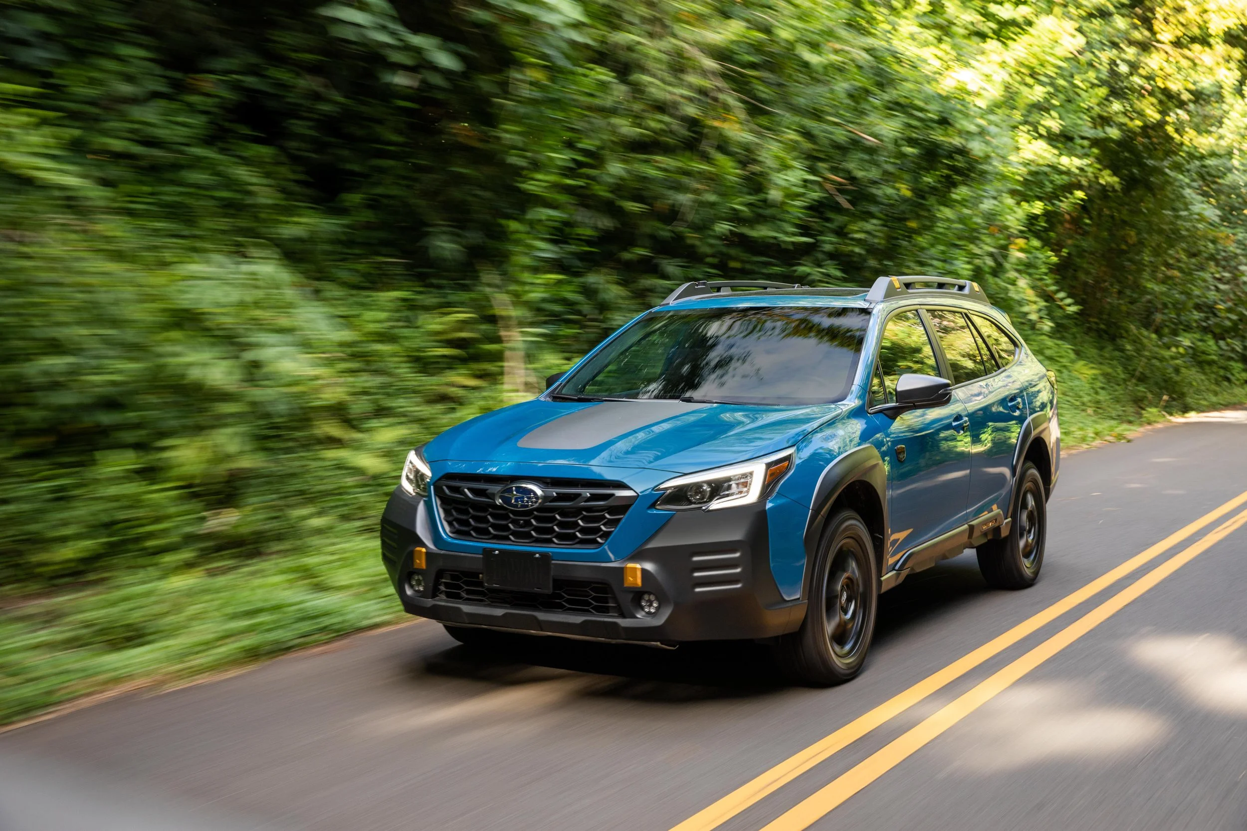 A blue Subaru Outback driving on a two-lane road surrounded by green trees and foliage, with a motion-blurred background indicating speed.