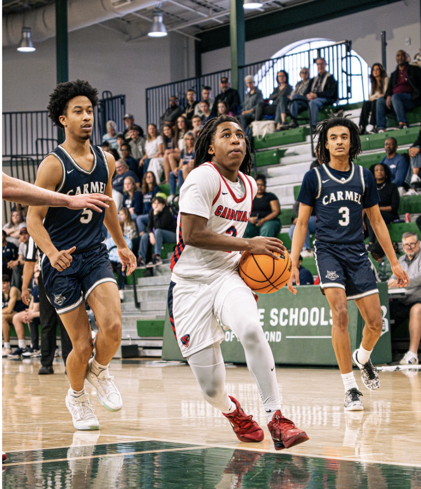 A basketball player in a white and red uniform dribbling the ball on the court, with two players in navy blue jerseys and an audience watching from the bleachers.