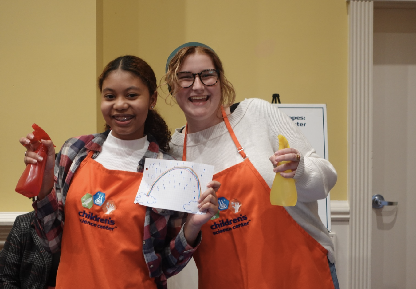 Two smiling girls holding spray bottles and a drawing of a rainbow rain scene at a children's science center event.
