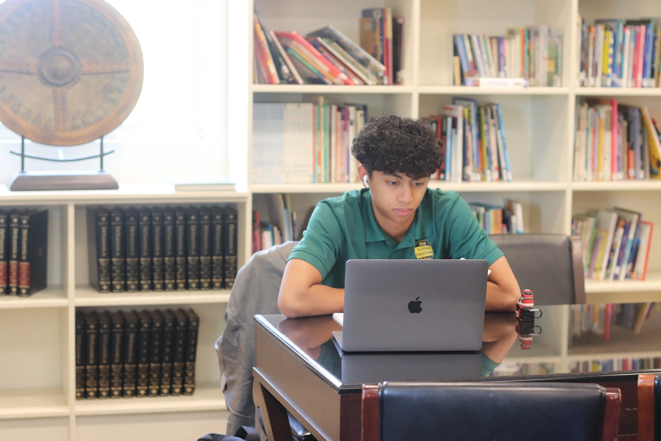 A teenage boy with curly hair sitting at a table working on a silver MacBook in a room with bookshelves and a globe.