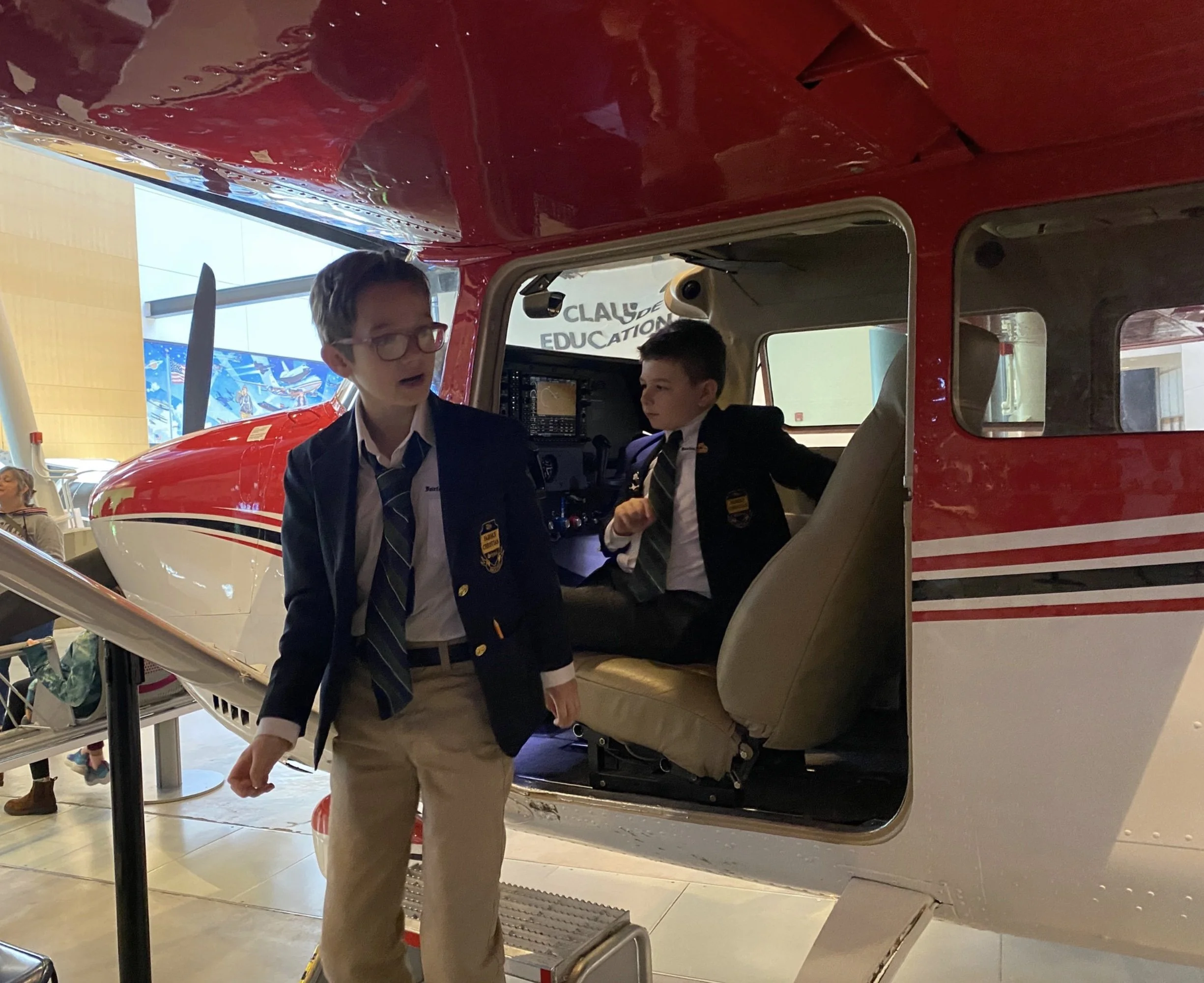 Two boys in school uniforms near a small red and white airplane displayed indoors, one standing outside and the other sitting inside the cockpit.