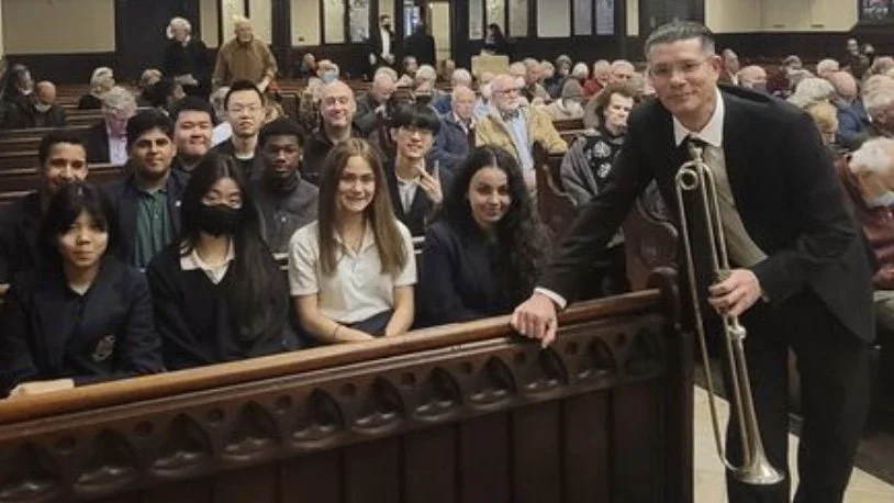 A group of young students sitting in a church or auditorium, some wearing face masks, with a man holding a trumpet standing beside them. The background shows many more seated people.