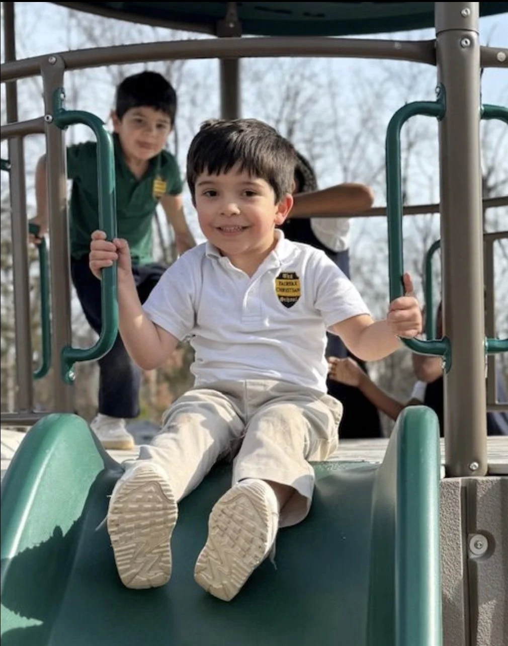 A young boy in a white school uniform sitting at the end of a playground slide, smiling and holding onto the metal bars, with other children in the background playing.