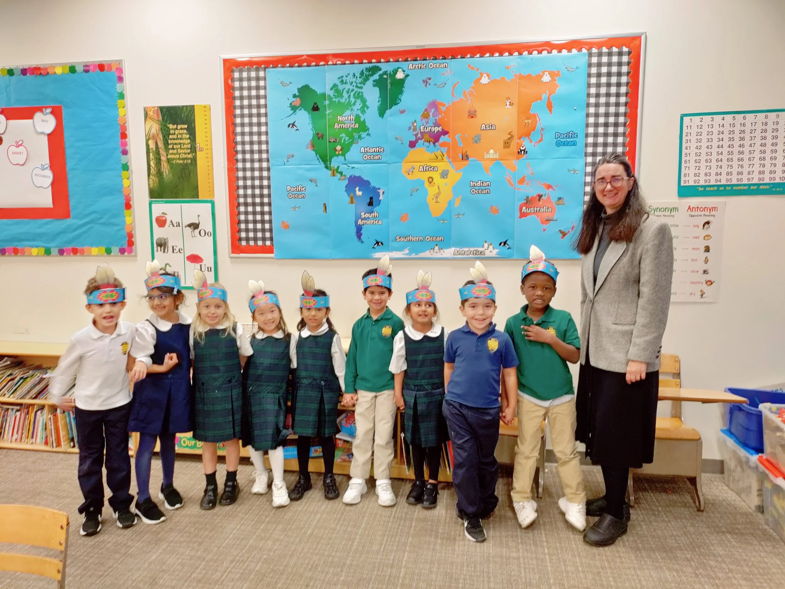 Group of young children wearing paper hats with bunny ears, standing in a classroom next to their teacher. The classroom has educational posters and a world map on the wall.