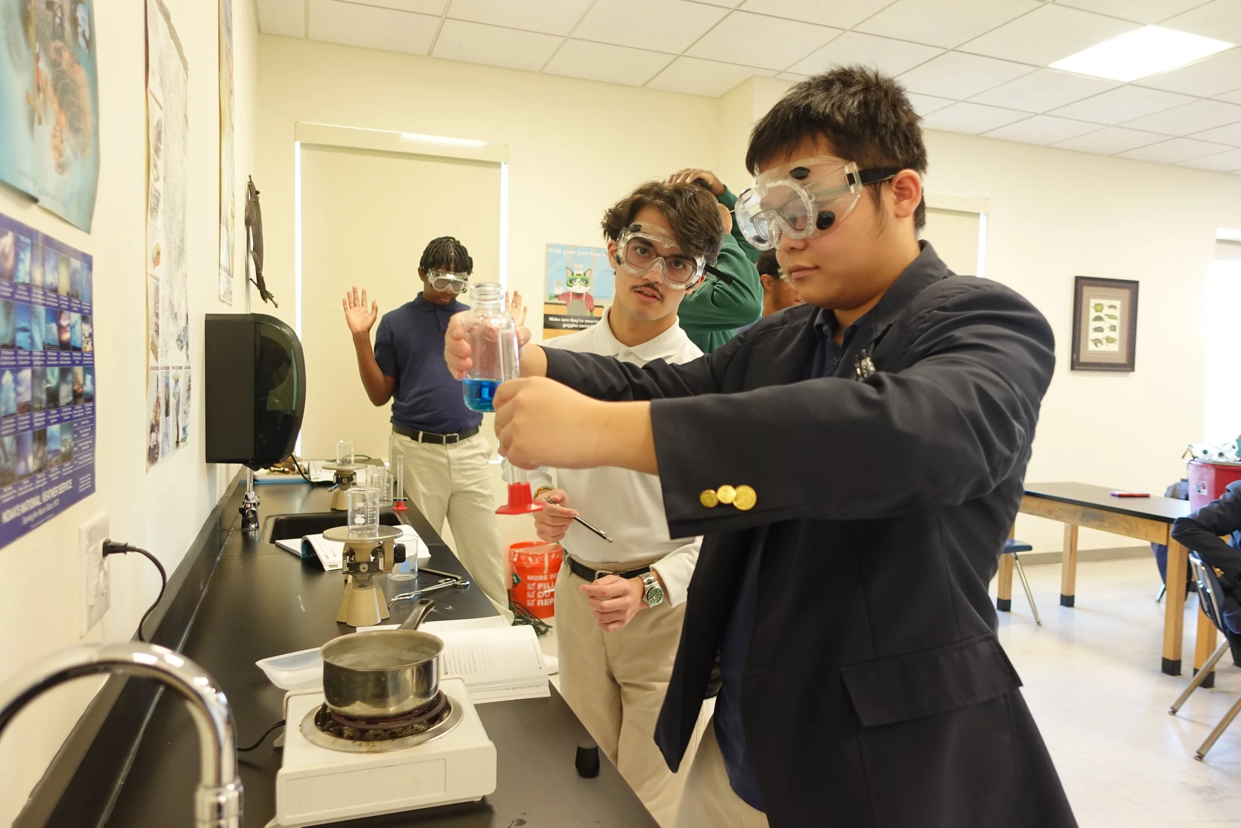 Students conducting a science experiment in a classroom, using safety goggles and handling a beaker with blue liquid.