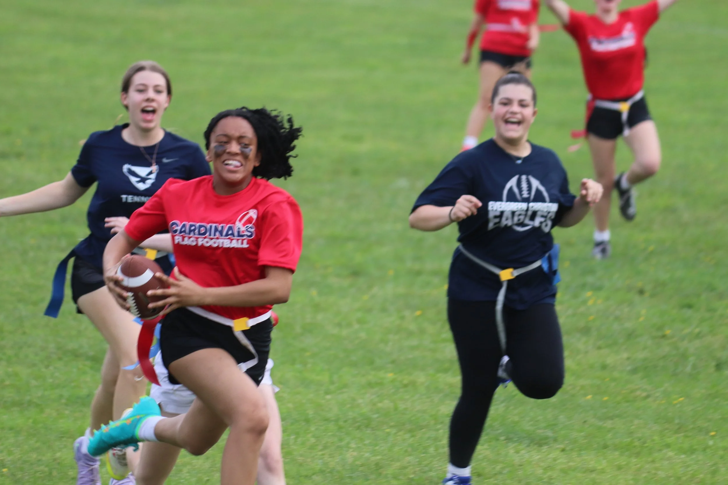 Young girls playing flag football outdoors on a grassy field, one girl in a red shirt holding a football and running while others chase her, all smiling and enjoying the game.