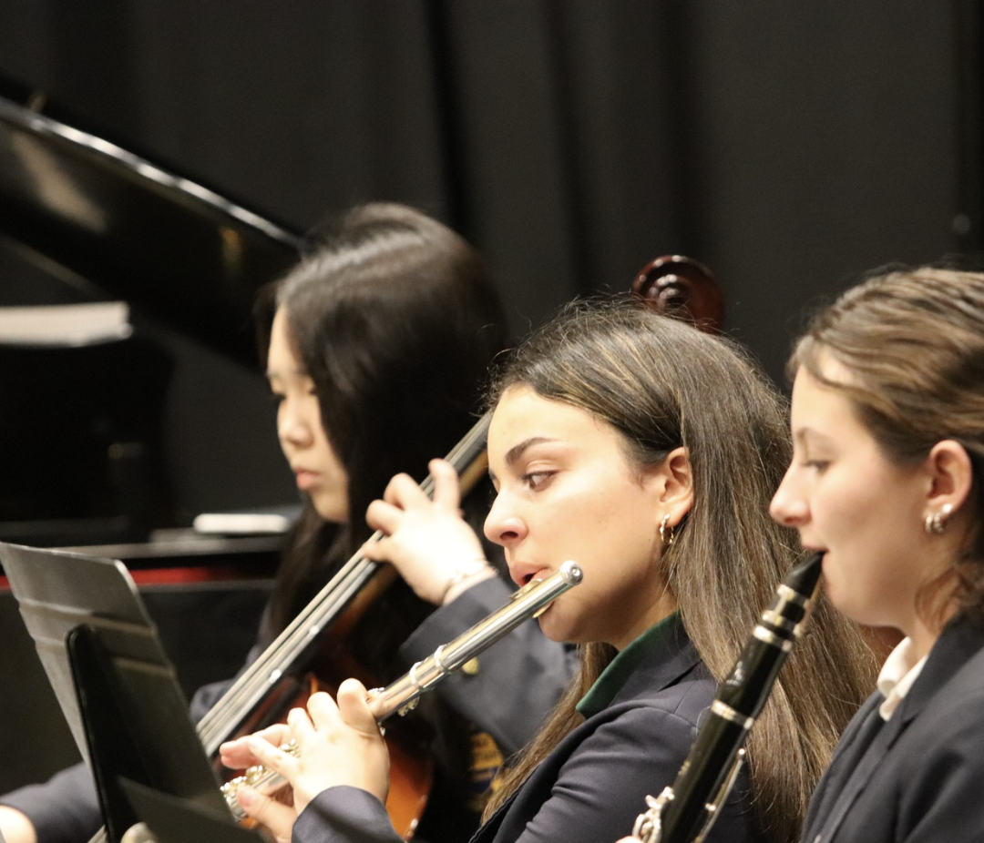 Three women playing musical instruments in an orchestra, with one playing the flute, one playing the violin, and one playing the clarinet.