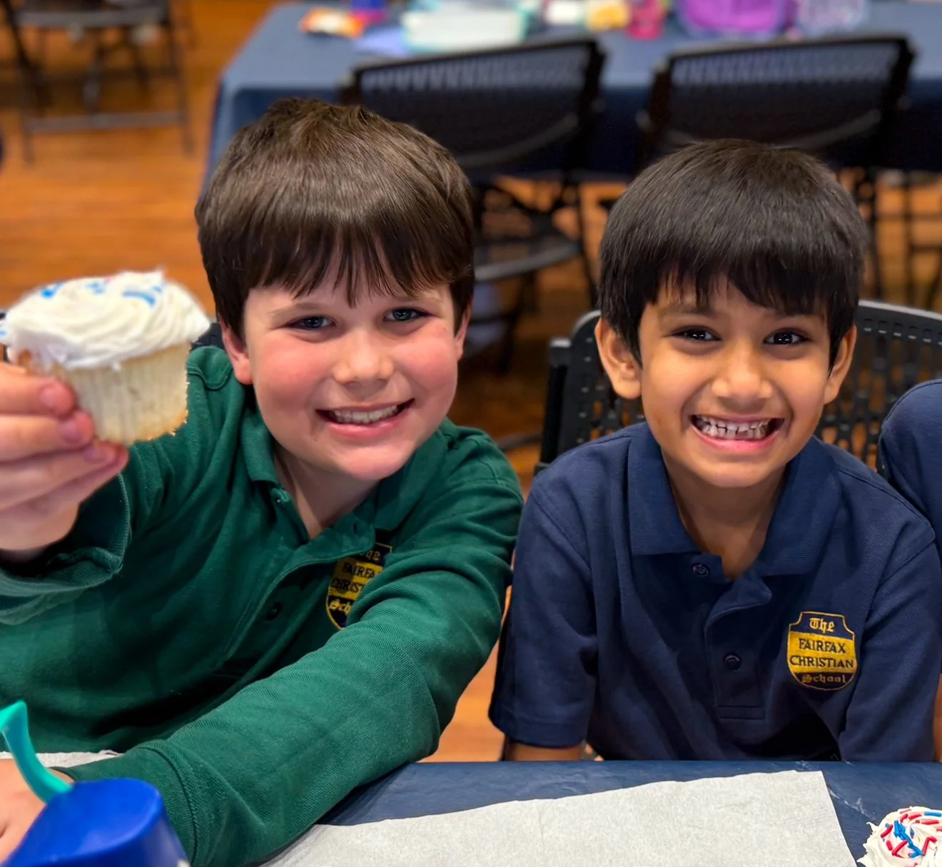 Two children smiling at a birthday celebration, one holding a cupcake with white frosting, sitting at a table with a paper napkin and a slice of cake nearby.