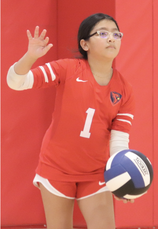 A young girl in a red volleyball jersey and shorts holding a volleyball, standing against a red background, raising her left hand.