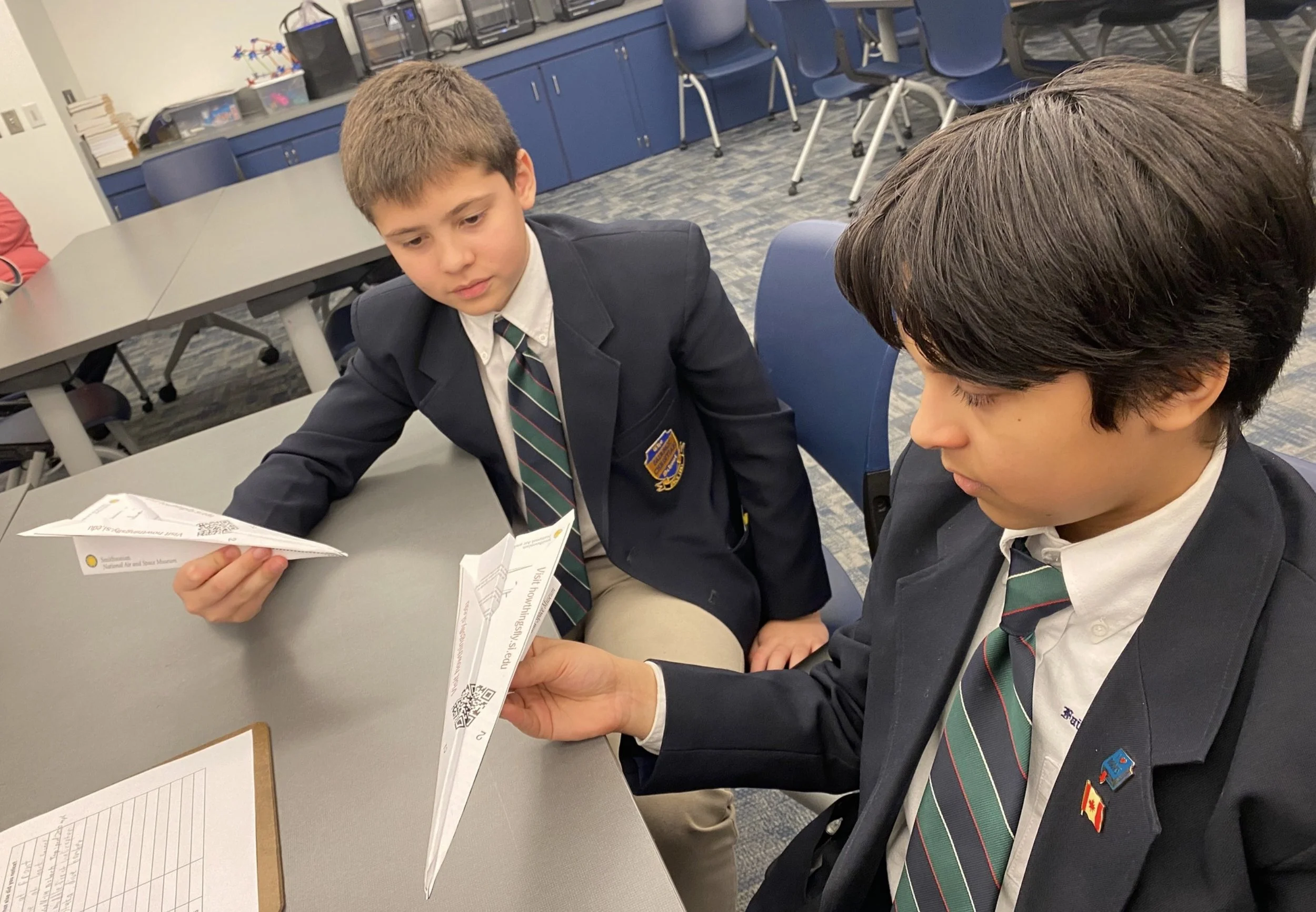 Two students in school uniforms sitting at a table, working with paper airplanes in a classroom or meeting room setting.