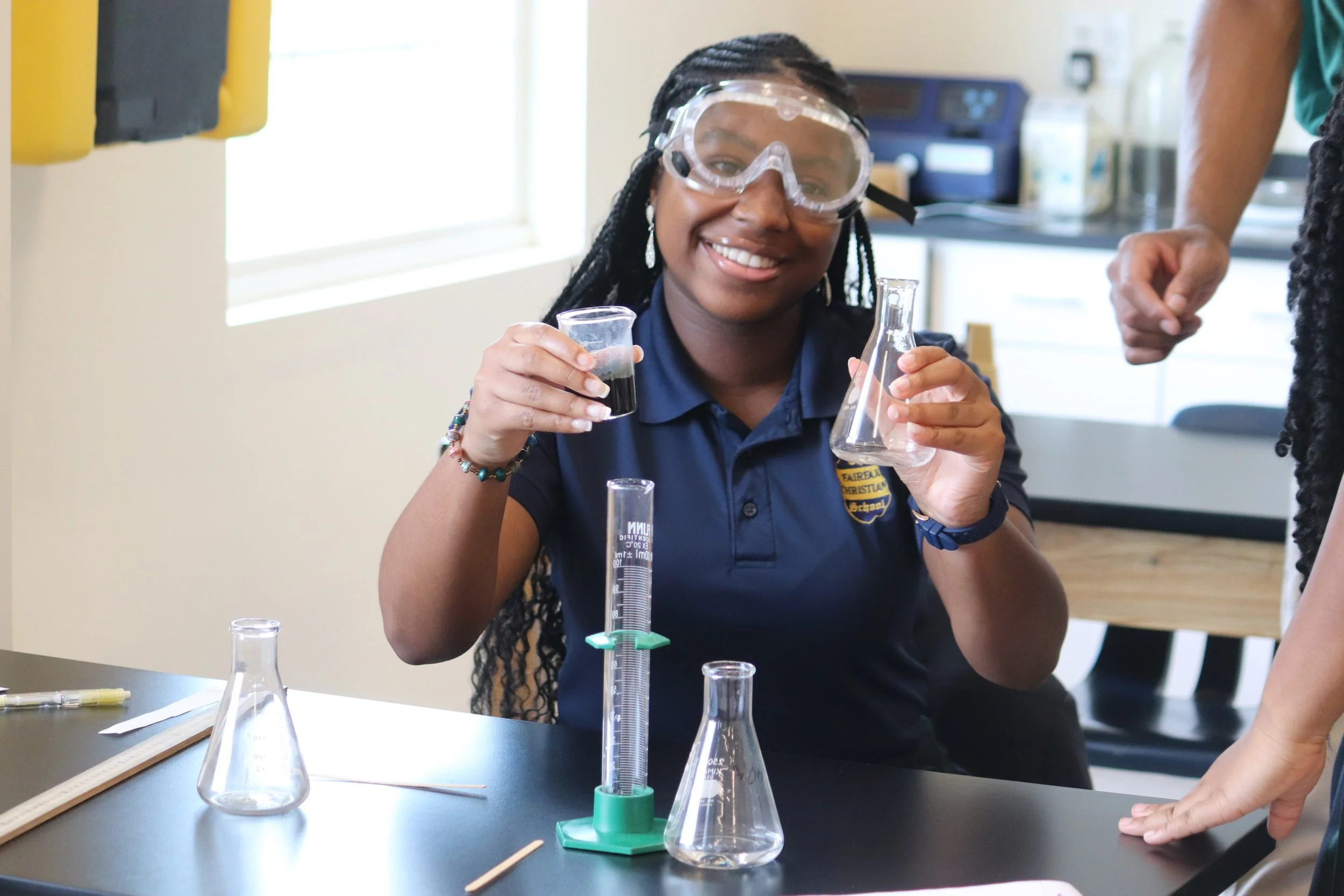 A young woman with glasses smiling while conducting a science experiment with laboratory glassware in a classroom.