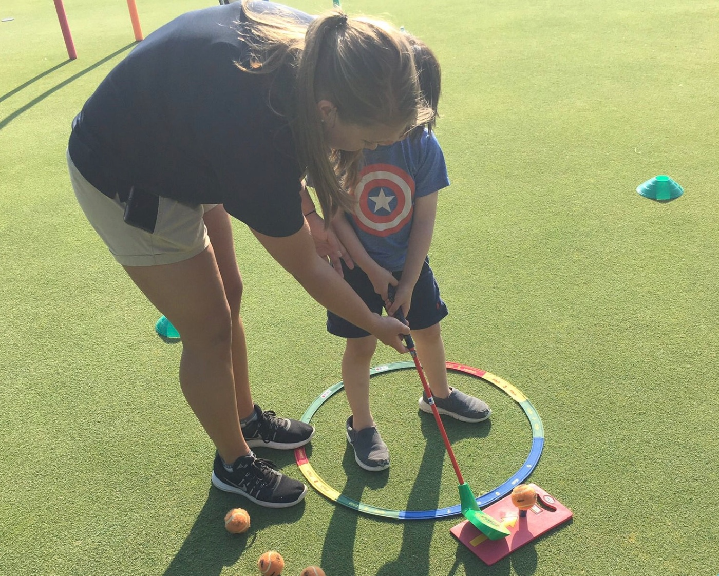A woman helping a young boy play mini-golf on a green course outdoors. The boy is holding a golf club aiming at an orange ball near a small white golf hole with pink and green barriers. The woman is guiding him, and there are several other orange balls on the ground nearby.