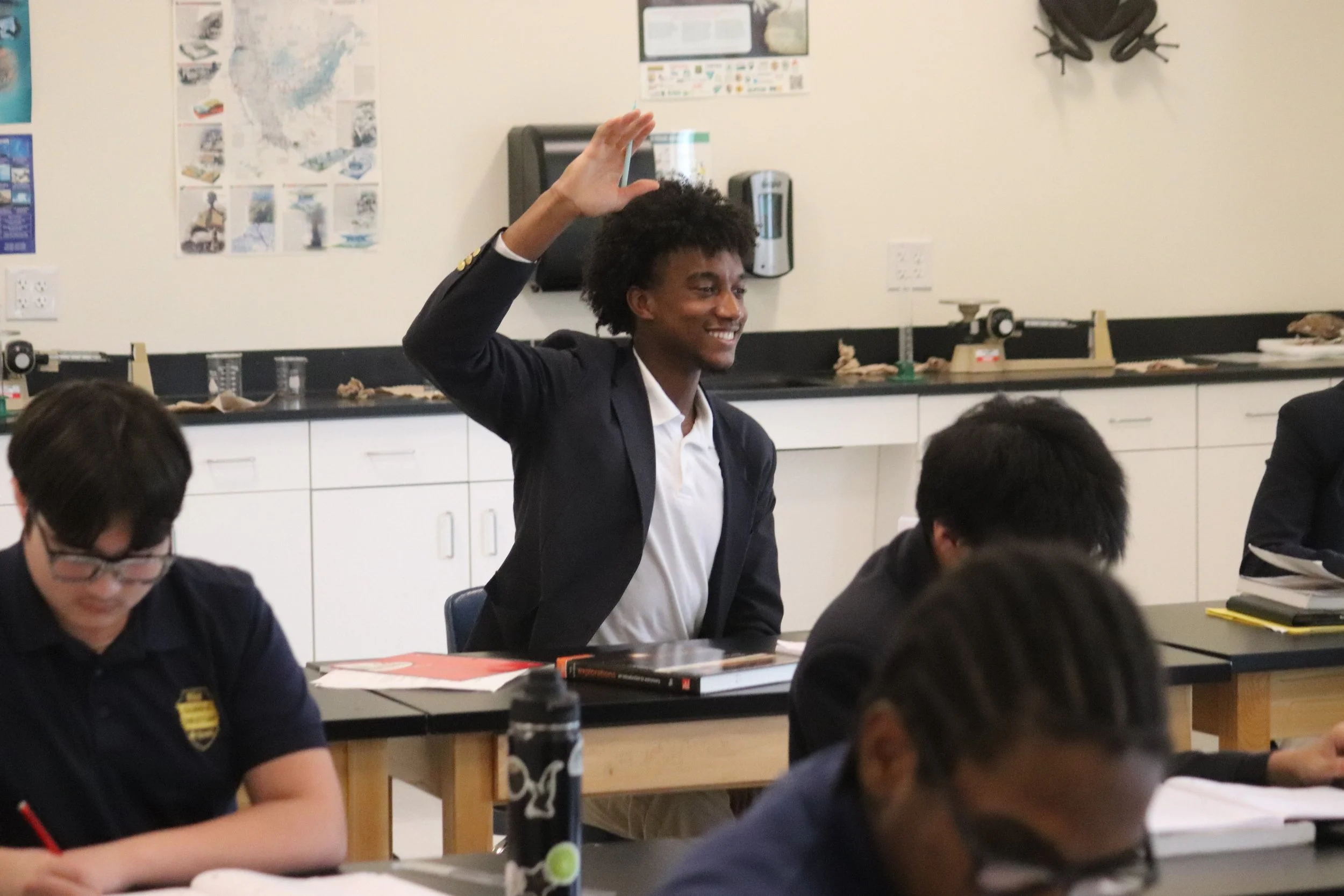 A young man in a suit sitting at a desk, smiling, raising his hand in a classroom with other students.