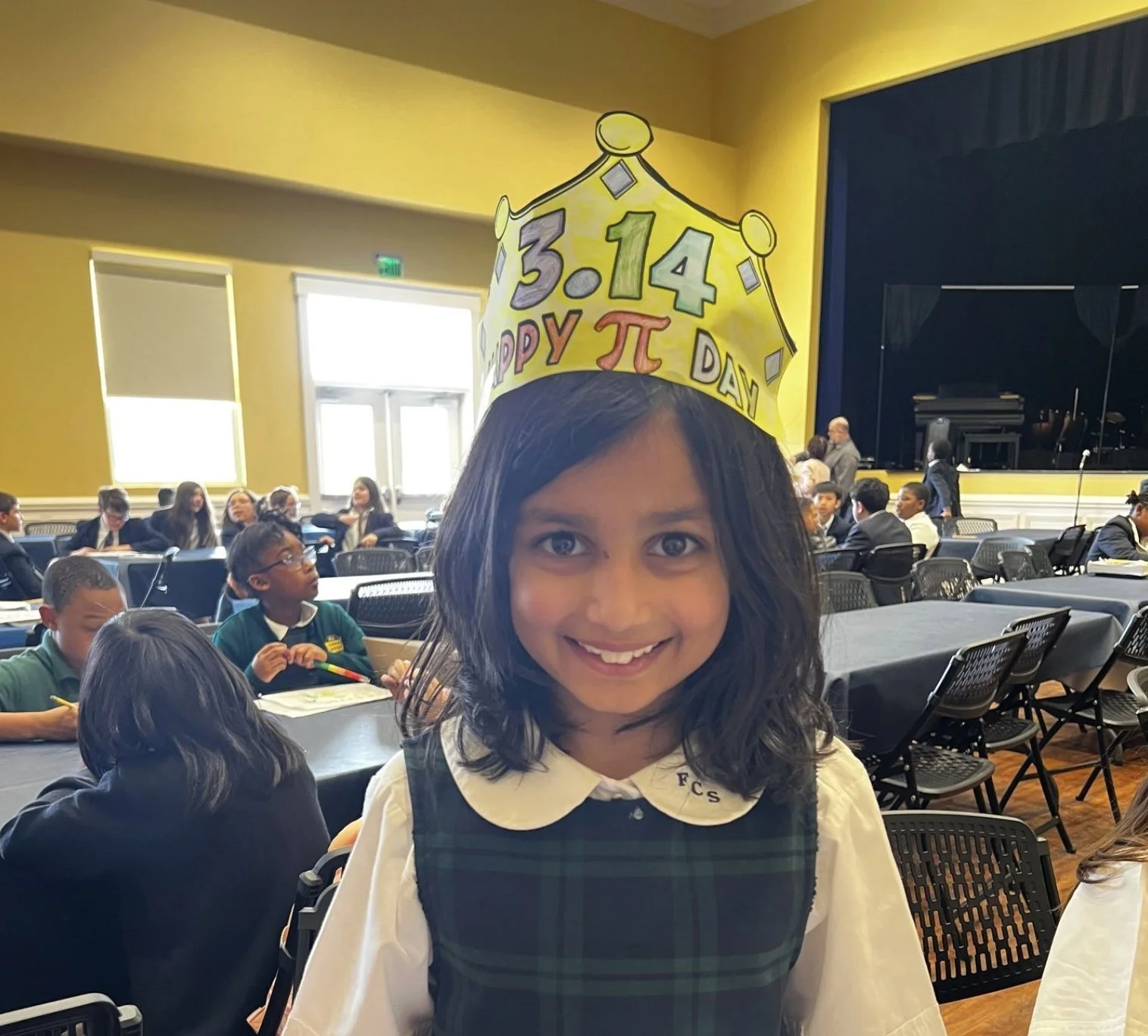 A young girl smiling at the camera wearing a yellow hat with the date 3.14 and 'Happy Pi Day' written on it, in a large hall with students seated at tables in the background.
