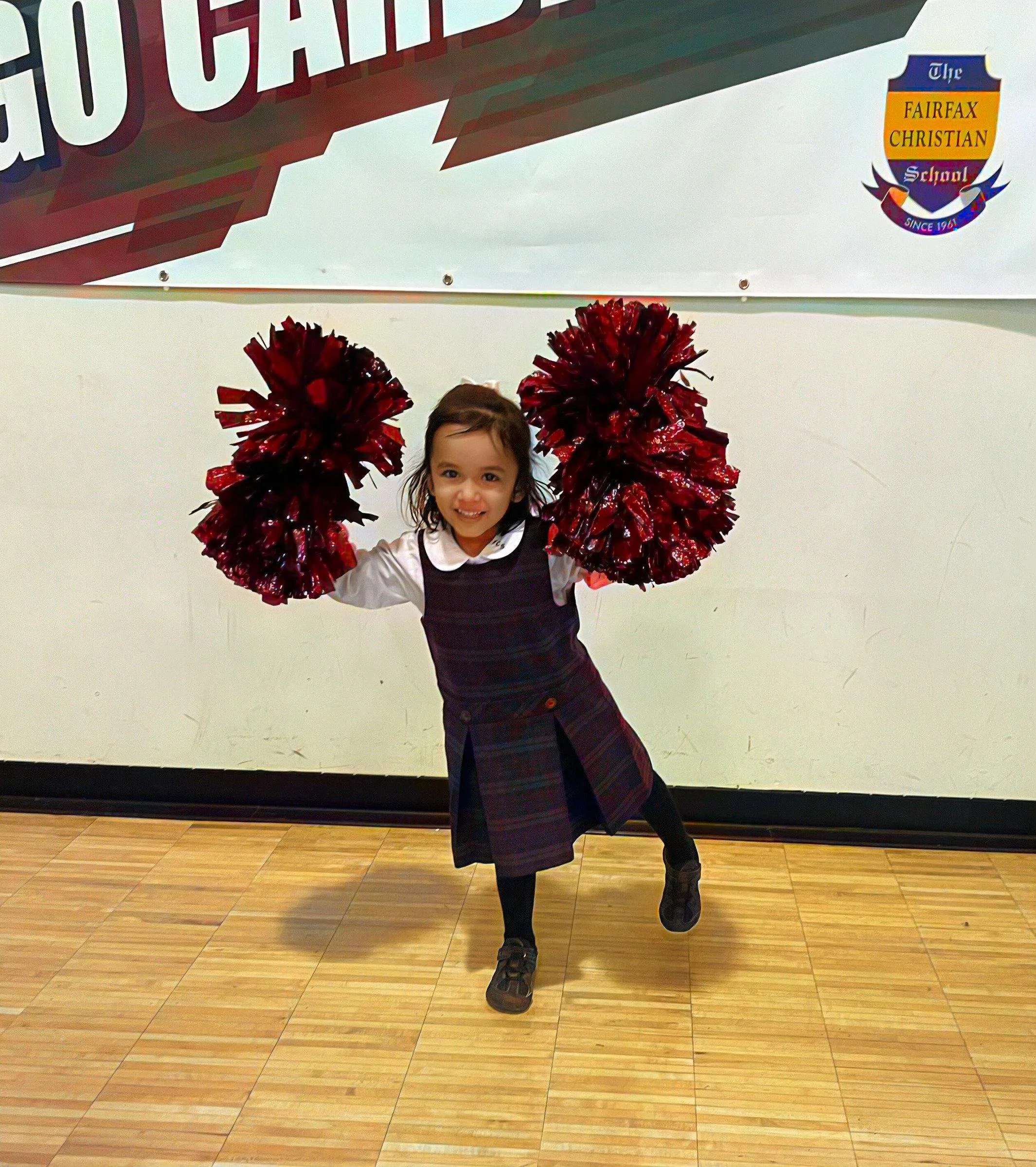 Young girl smiling and holding red pom-poms, standing in front of a school banner with a Fairfax Christian School logo.