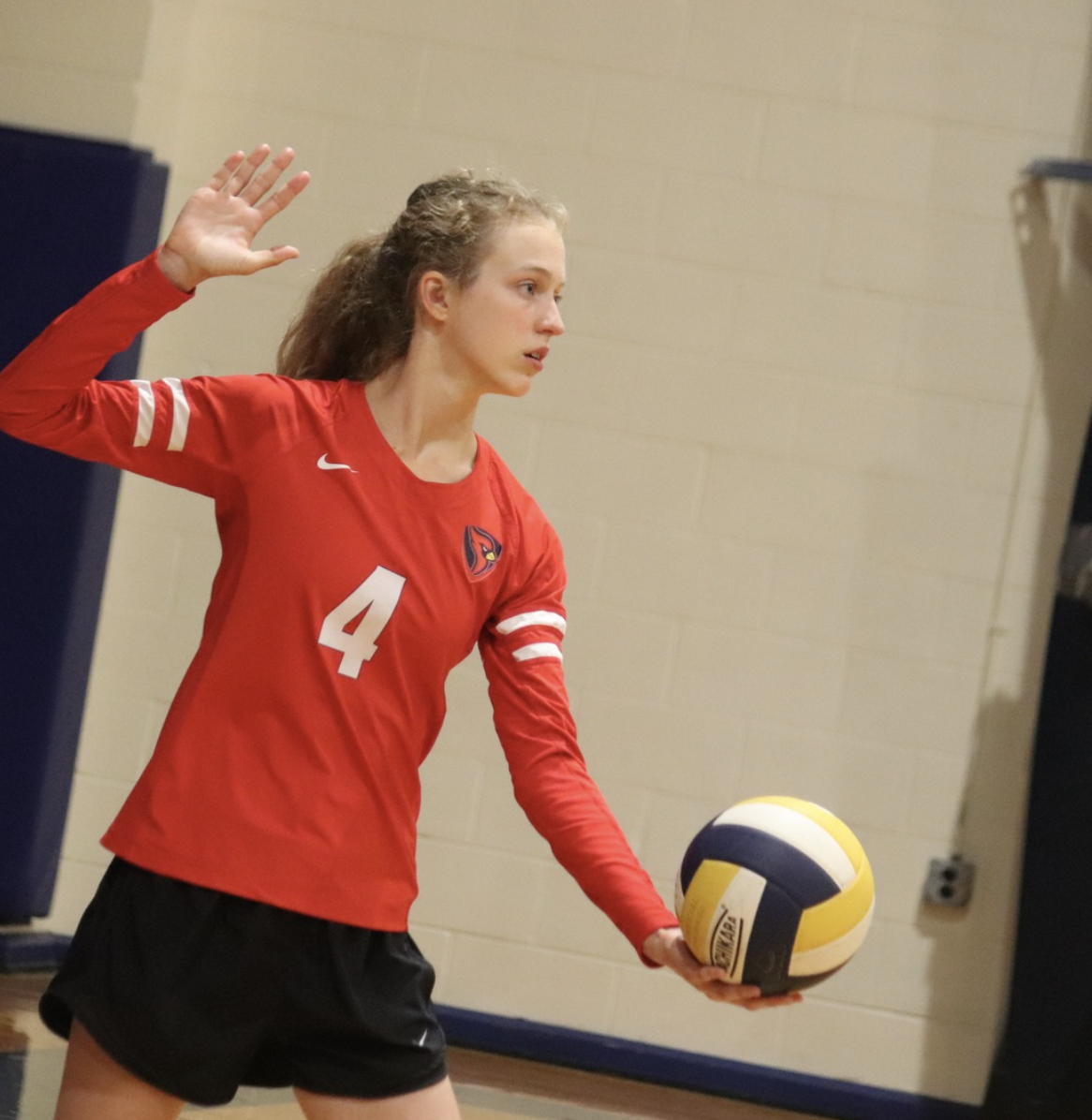A young female volleyball player holding a volleyball, wearing a red jersey with the number 4, and preparing to serve during a game in an indoor gym.