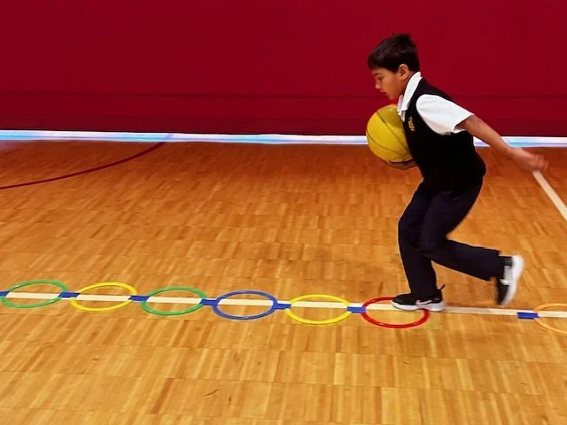 A boy in school uniform jumping over colorful hoops on a wooden gym floor while holding a yellow basketball.
